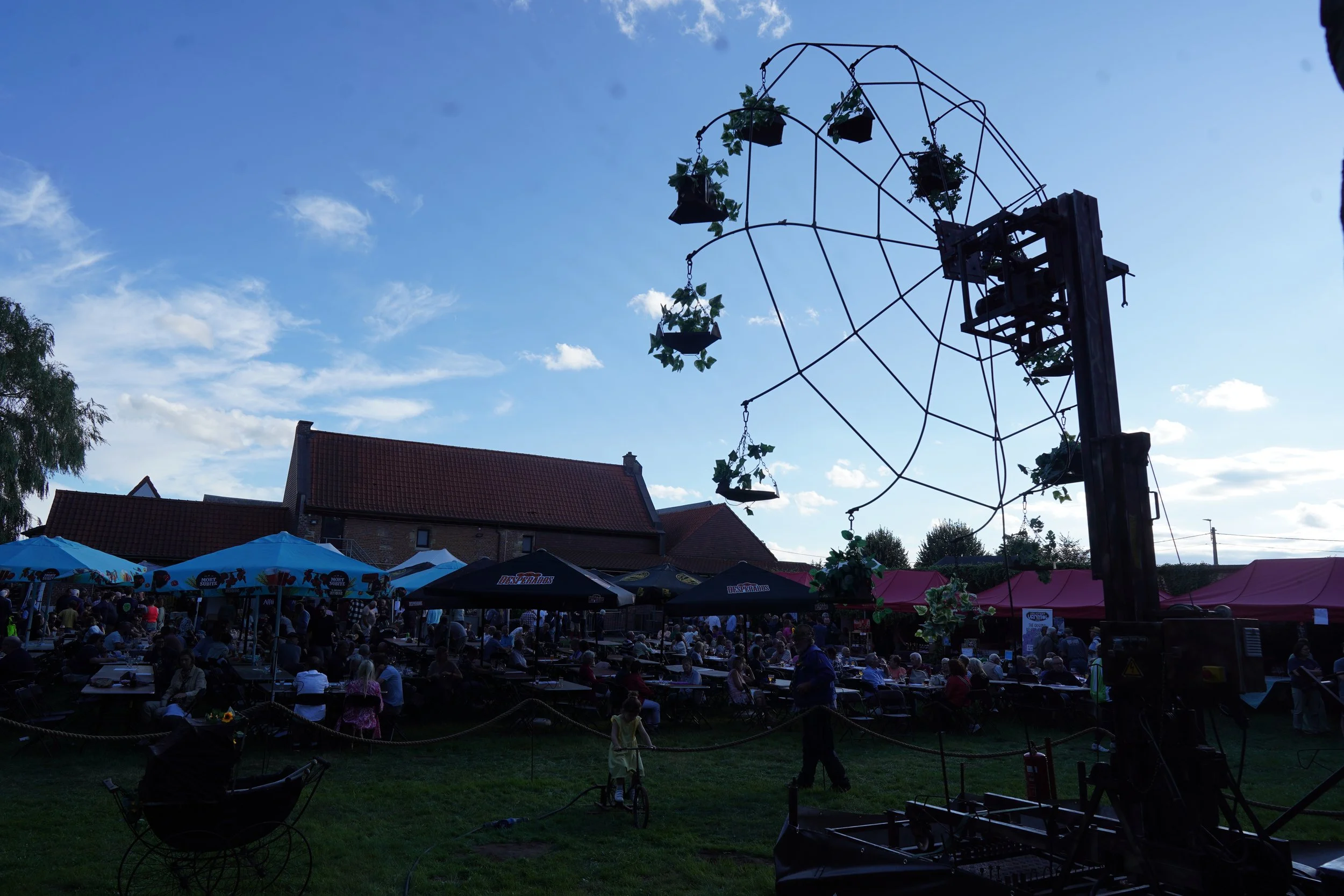 Een avondmarkt met mensen die aan tafels eten, onder parasols, met een grote antieke schompferris in de achtergrond en een kinderfietser op het gras. Bellenmolen, vuurmolen, onthaal, vuuranimatie, vuurspuwer, vuurartiest, vuurshow.