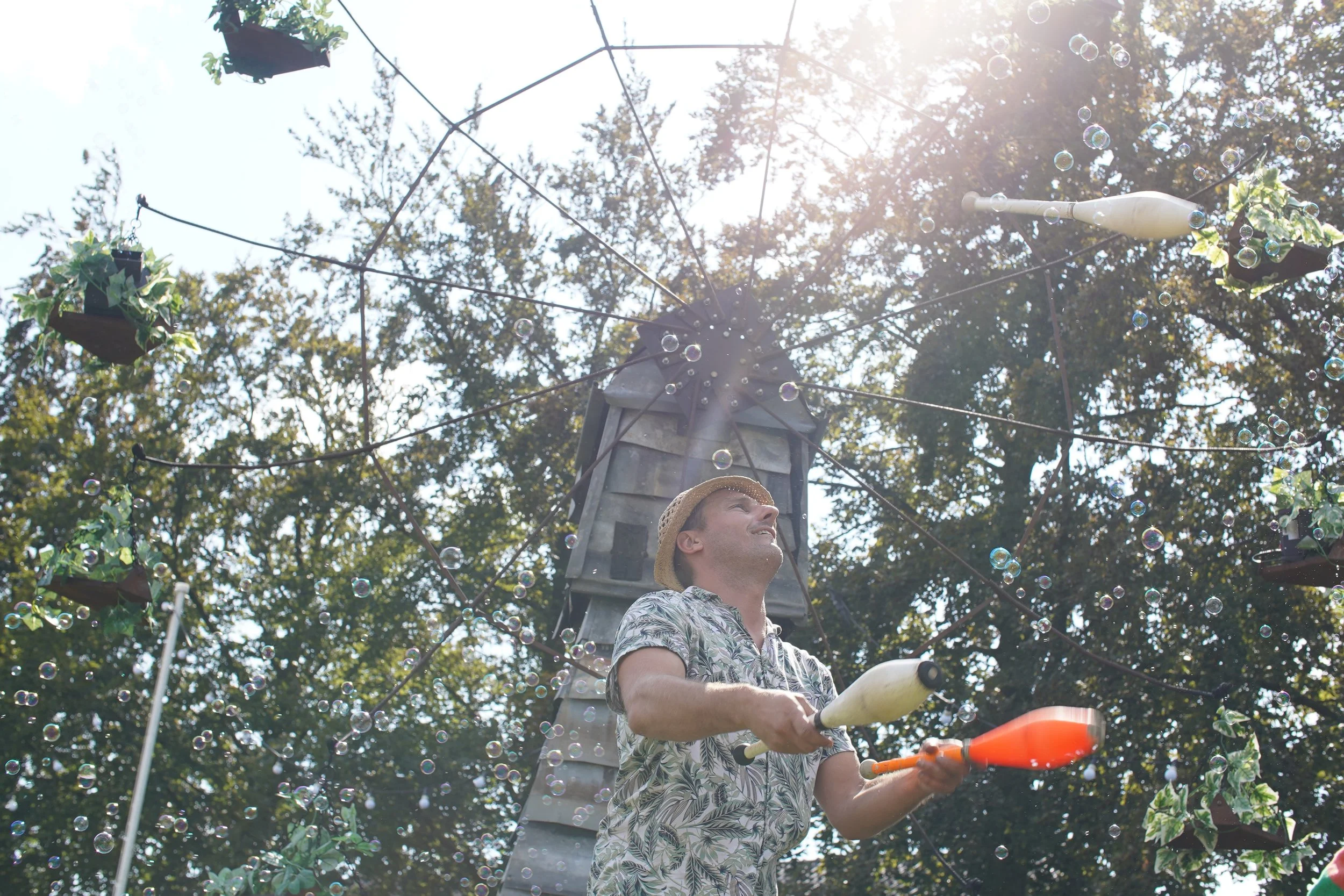 Man in een Hawaii-hemd en hoed die met ballonnen in de hand speelt op een buitenspeelplek met een toren en bomen, zon en zeepbellen. Bellenmolen, vuurmolen, onthaal, vuuranimatie, vuurspuwer, vuurartiest, vuurshow.