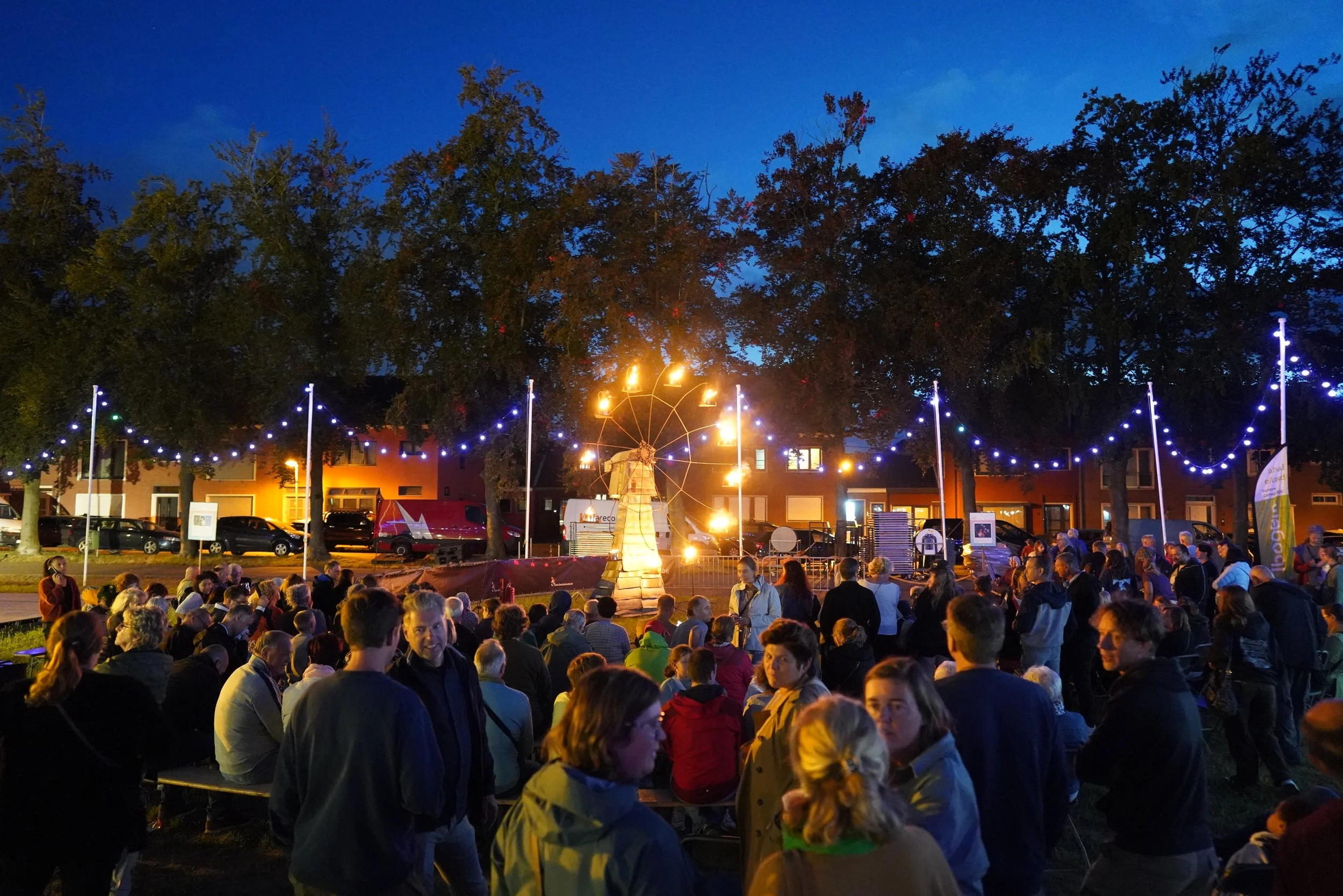 Een avondbijeenkomst in een park met een grote groep mensen die naar een voorstelling of evenement kijken, met verlichting en een draaiende wiek in het midden, onder een blauwe hemel en omgeven door bomen.