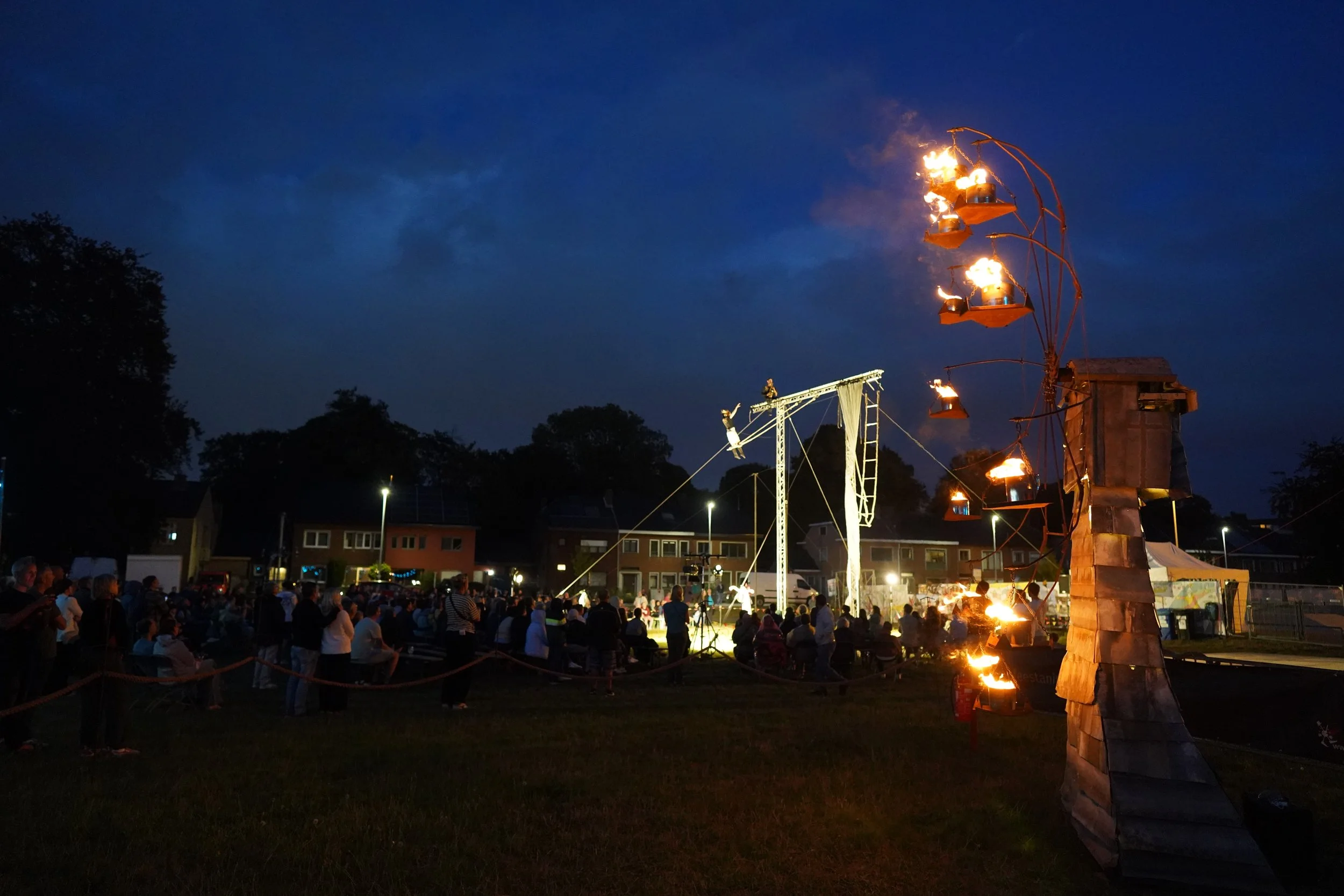 Mensen kijken naar een vuurshow met vlammen en een draaiende constructie in een openluchtfeest bij nacht, met huizen op de achtergrond. Bellenmolen, vuurmolen, onthaal, vuuranimatie, vuurspuwer, vuurartiest, vuurshow.