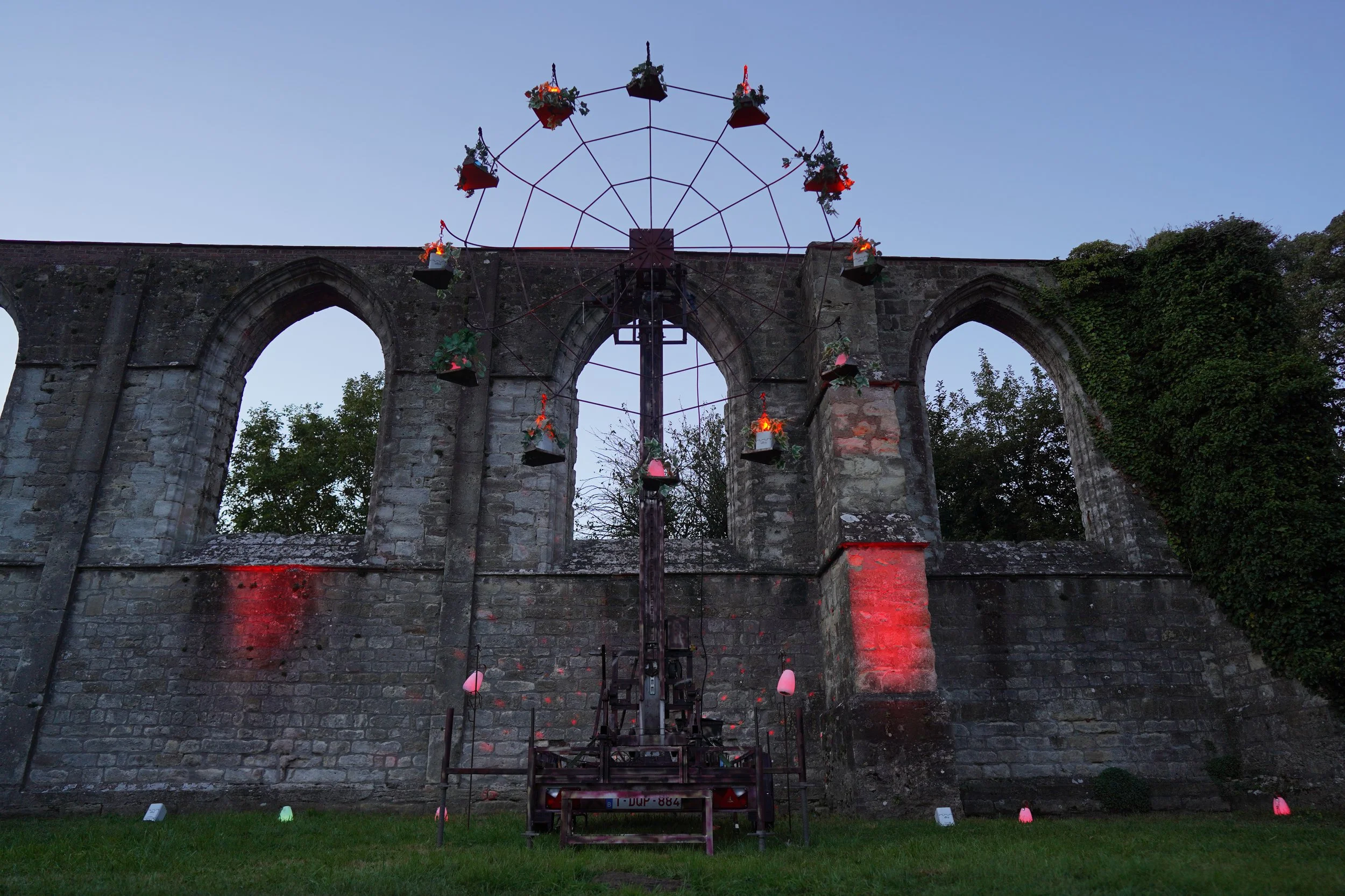 Een oude stenen muur met drie grote boogvensters, bedekt met groene klimop aan de rechterkant. Voor de muur staat een klein, metalen wiel met planten en lampjes, de lichtmolen. Bellenmolen, vuurmolen, onthaal, vuuranimatie, vuurspuwer, vuurartiest.