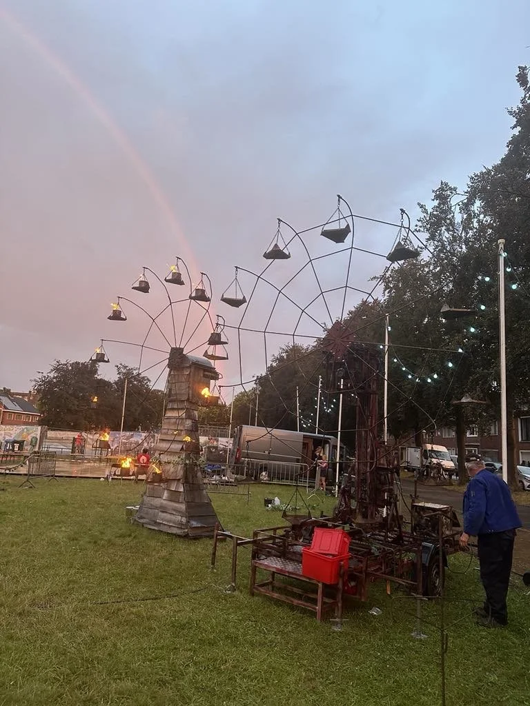 Een reuzenrad met kleine gondels in een park, met een schemerige lucht en een regenboog op de achtergrond. Mensen staan erbij, en er zijn bomen en trucks. Bellenmolen, vuurmolen, onthaal, vuuranimatie, vuurspuwer, vuurartiest, vuurshow.