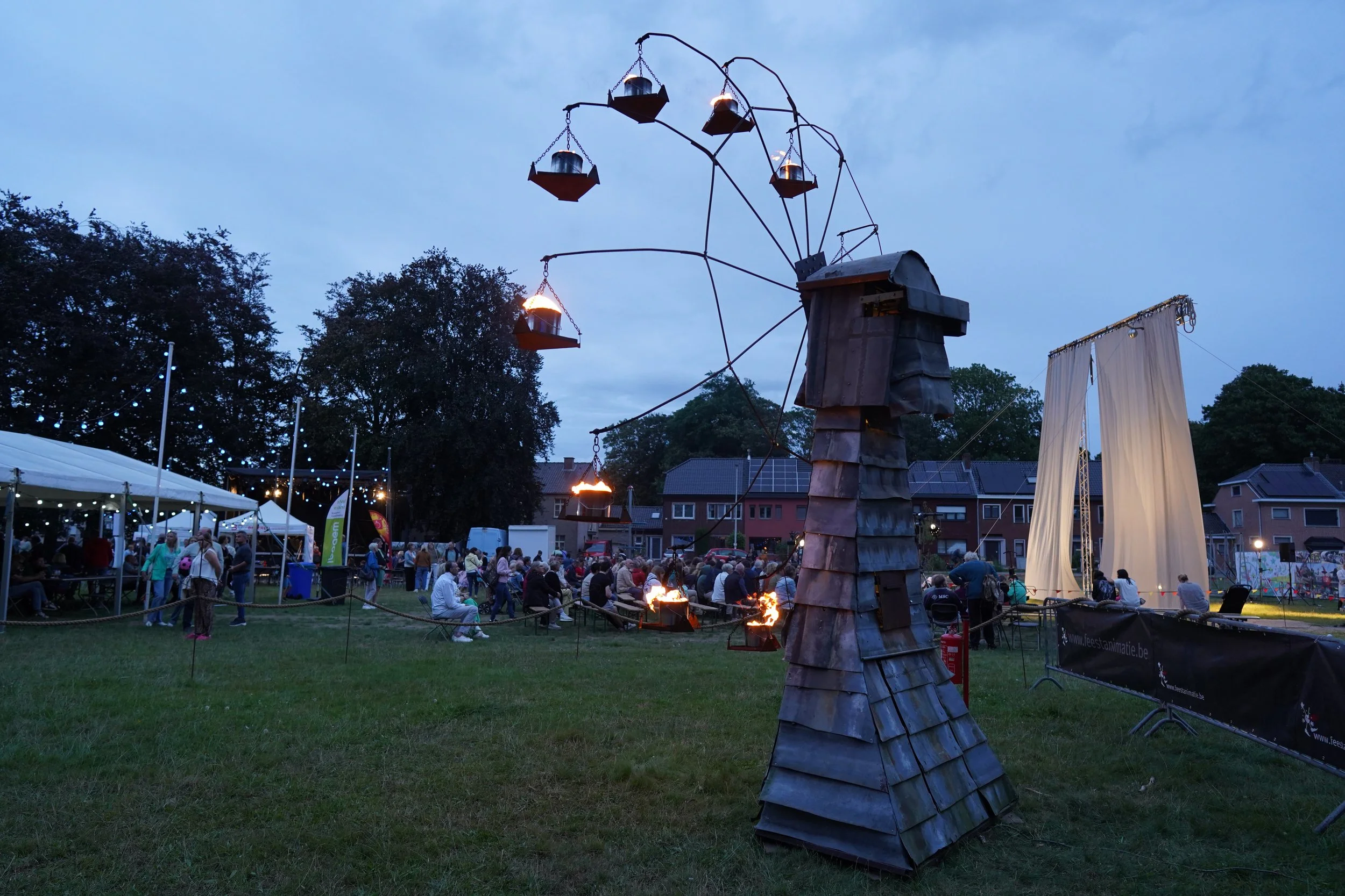 Een openlucht evenement met mensen die toeschouwers zitten op stoelen en staan op gras, terwijl er een kunstwerk met brandende lampen op de voorgrond staat, onder een blauwe bewolkte hemel.