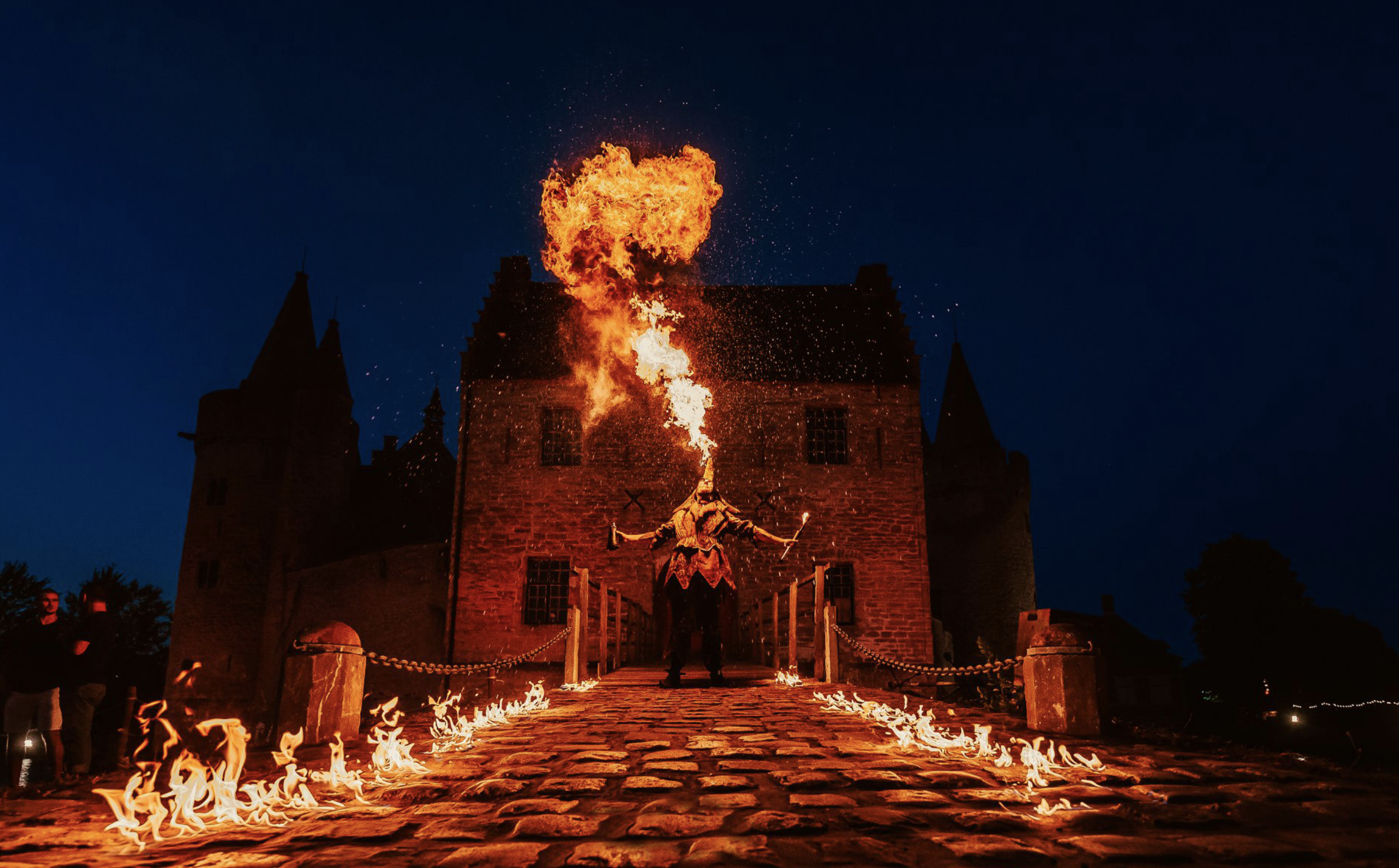 Een vuurspuwende performer op een kasteelbrug tijdens de nacht, met vuur rondom en op de grond, en een donkerblauwe hemel op de achtergrond. Bellenmolen, vuurmolen, onthaal, vuuranimatie, vuurspuwer, vuurartiest, vuurshow.