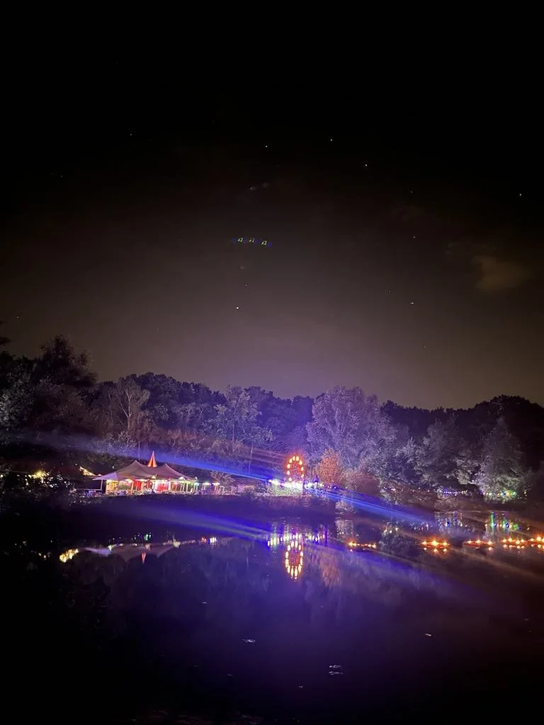 Een nachtelijk scène met een meer dat weerspiegelt, een verlichte kermis of festival met een tent en een reuzenrad, omgeven door bomen en een heldere donkere hemel met sterren. Bellenmolen, vuurmolen, onthaal, vuuranimatie, vuurspuwer, vuurartiest.