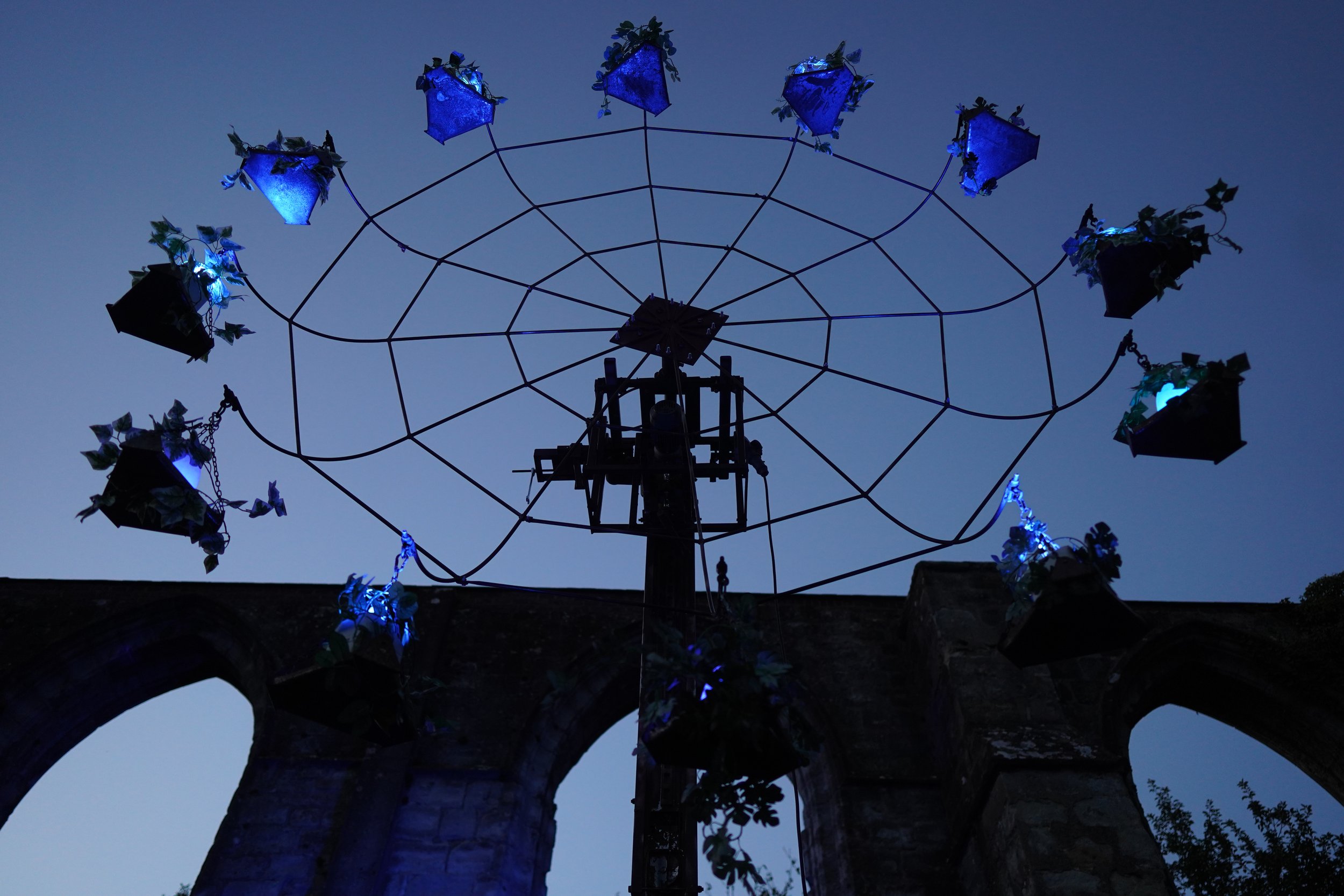 Uitzicht vanaf de grond op een oud wieltje met kunstlicht, bloemen en bladeren, tegen een donkerblauwe avondlucht. Bellenmolen, vuurmolen, onthaal, vuuranimatie, vuurspuwer, vuurartiest, vuurshow.