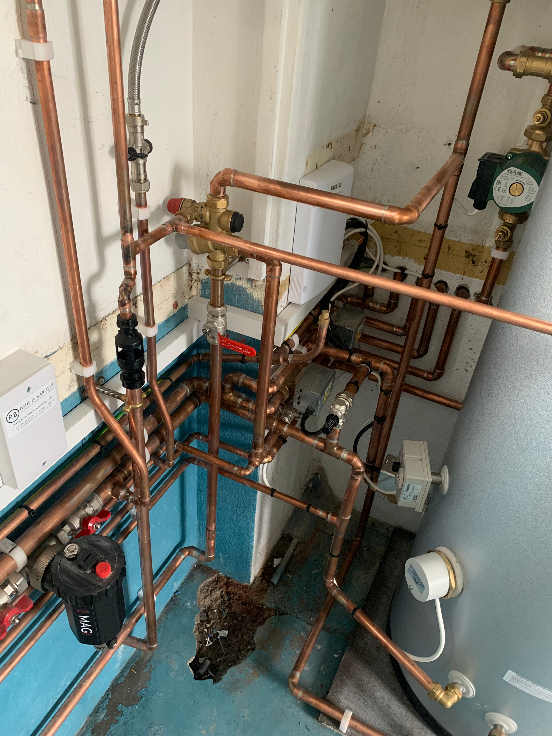 Close-up view of plumbing pipes, valves, and equipment in a utility room, with a hole in the floor and exposed pipes.