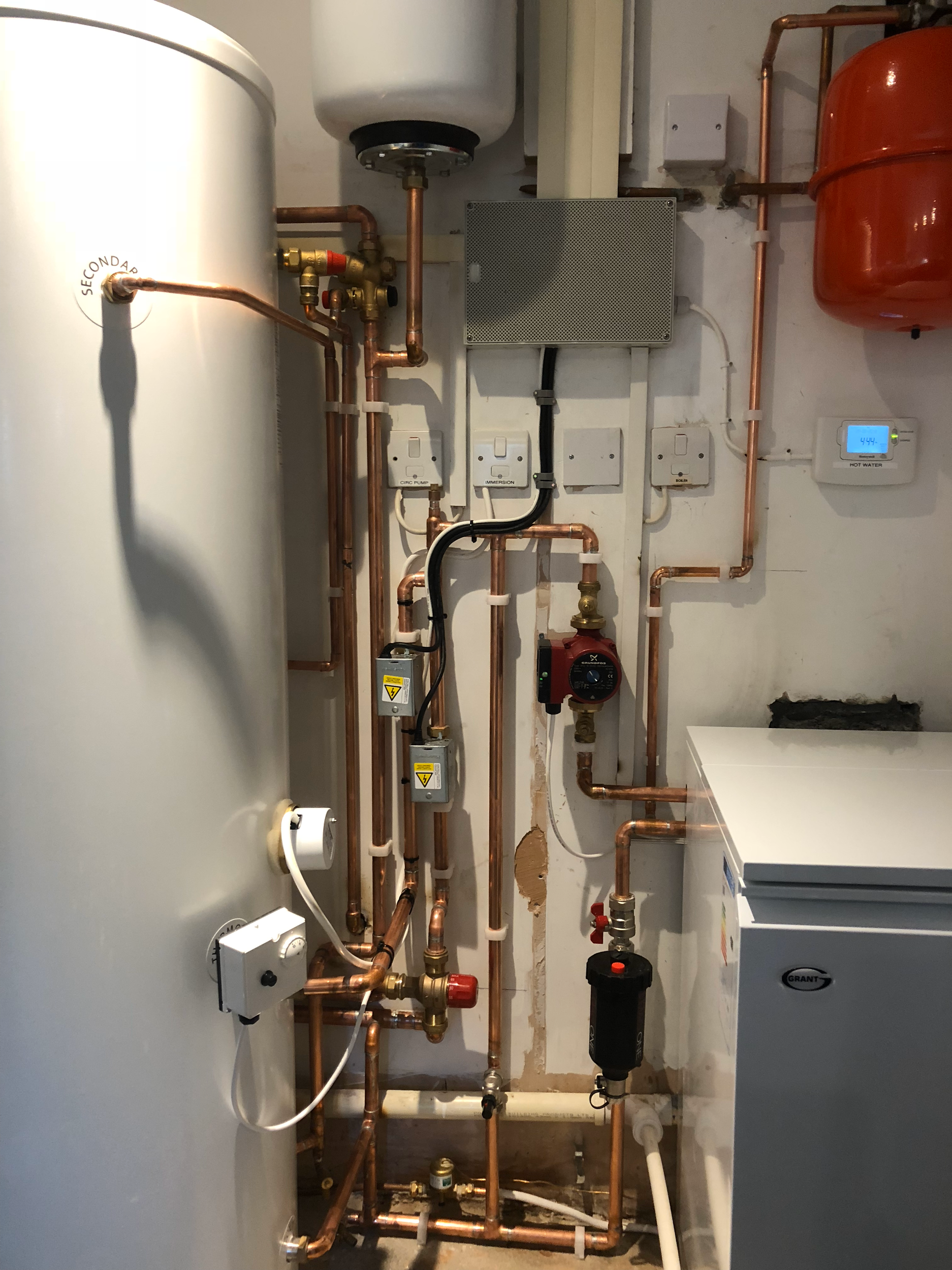 A boiler room with a large white water heater tank on the left and copper pipes, gauges, and electrical components on the wall.
