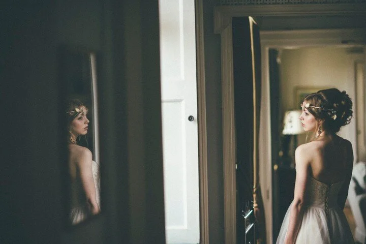 A bride glances out of the bedroom window at her groom and guests waiting on the lawn below