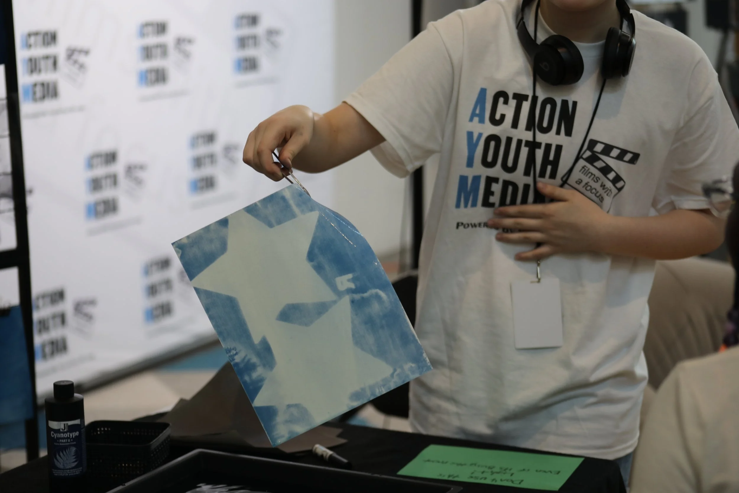 A piece of paper being held with tongs. The paper is wet, appearing to be a cyanotype photo depicting two white stars and the letter R. The letter R is reversed.