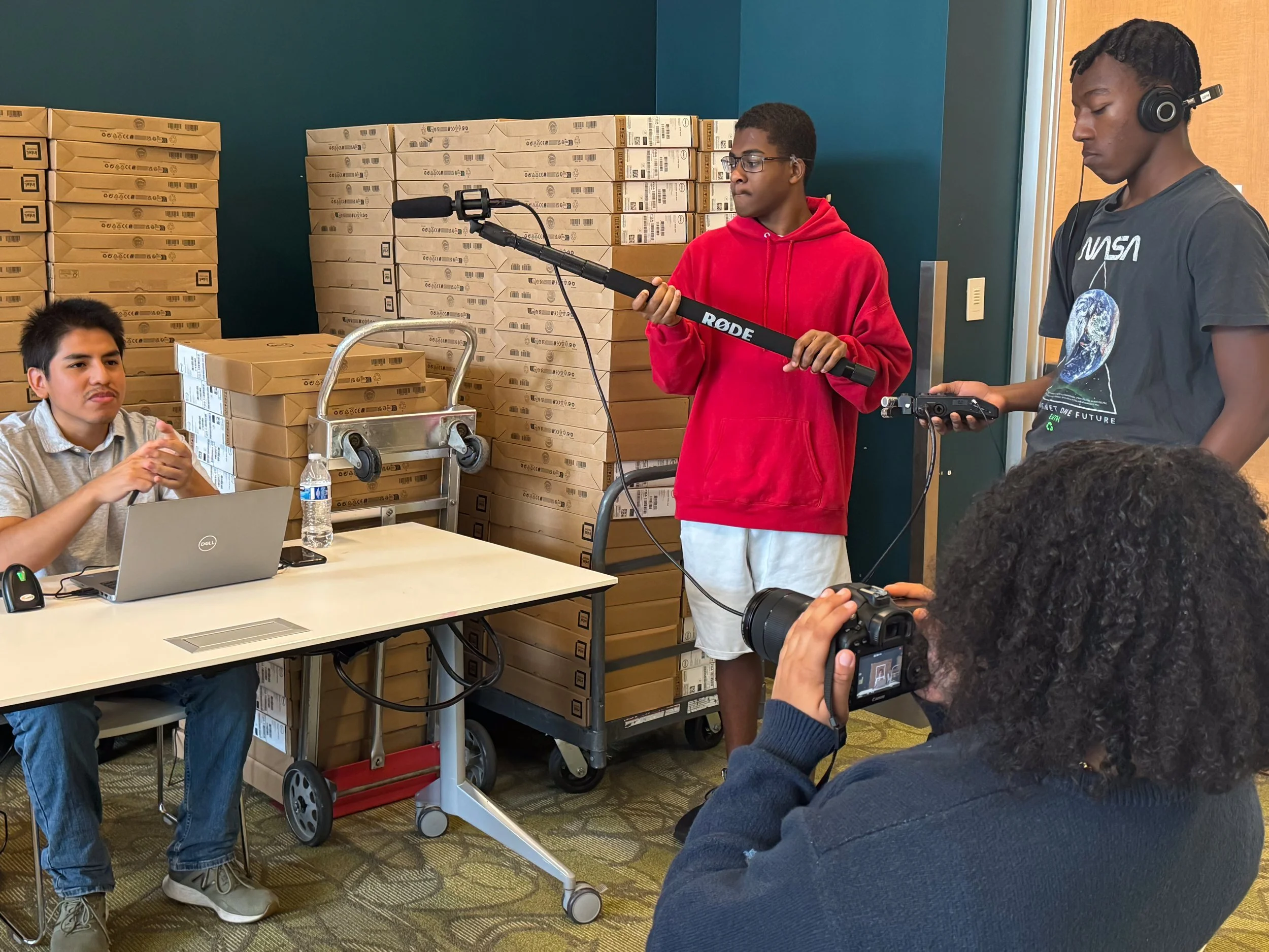 A person sits at a table with a laptop. He is in front of stacks of boxes, presumably containing laptops. Three people are around him. One holds a boom mic, another is recording audio, and the third is recording with a camera.