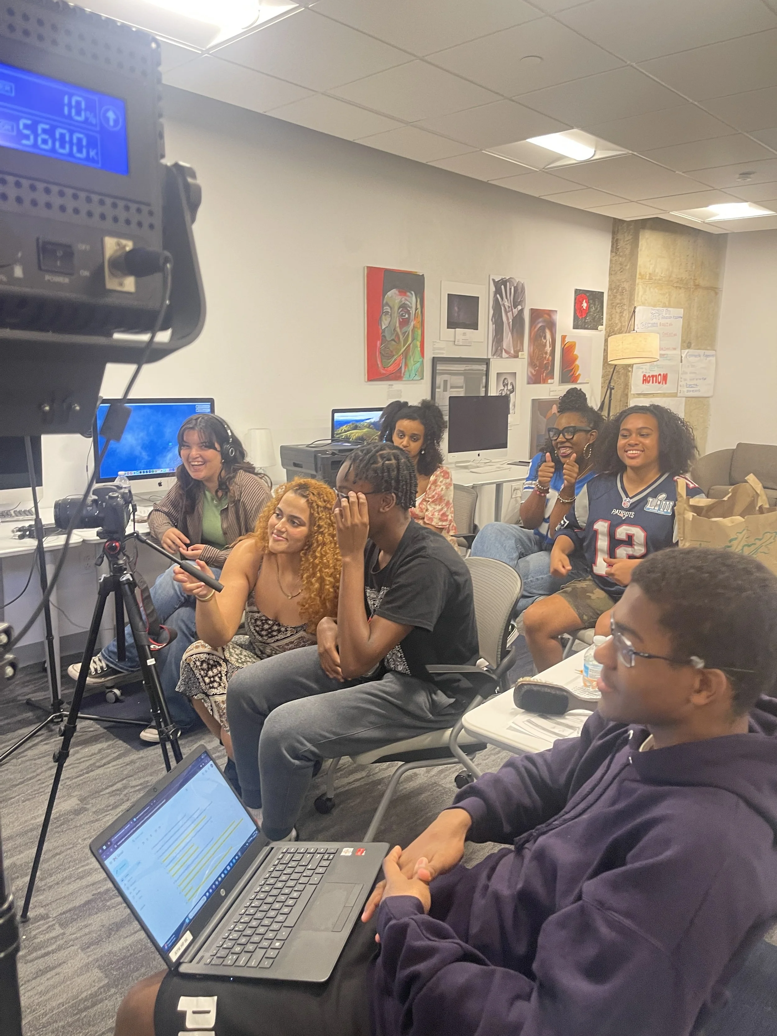 Seven people sitting, looking towards the left at something out of frame. One person in the front has a laptop open, one in the back is operating a camera, and standup studio lights are pointed in the direction they are facing.