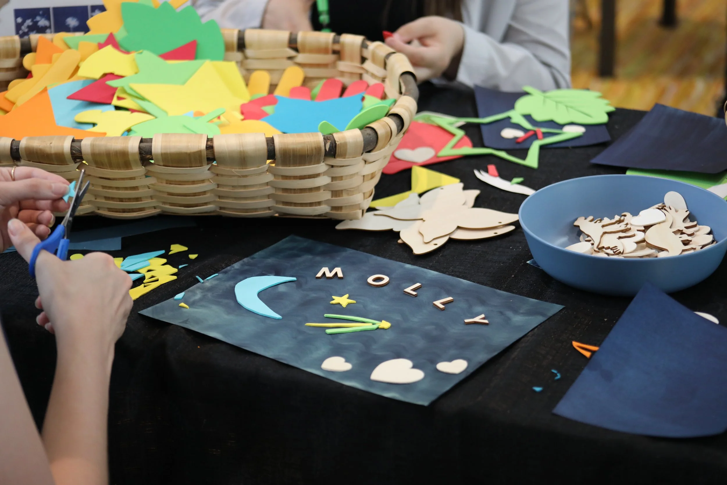A painted sheet of paper covered in brightly colored foam shapes and wooden cutouts. This is done to create a cyanotype photo. To the left is a person cutting a foam star.