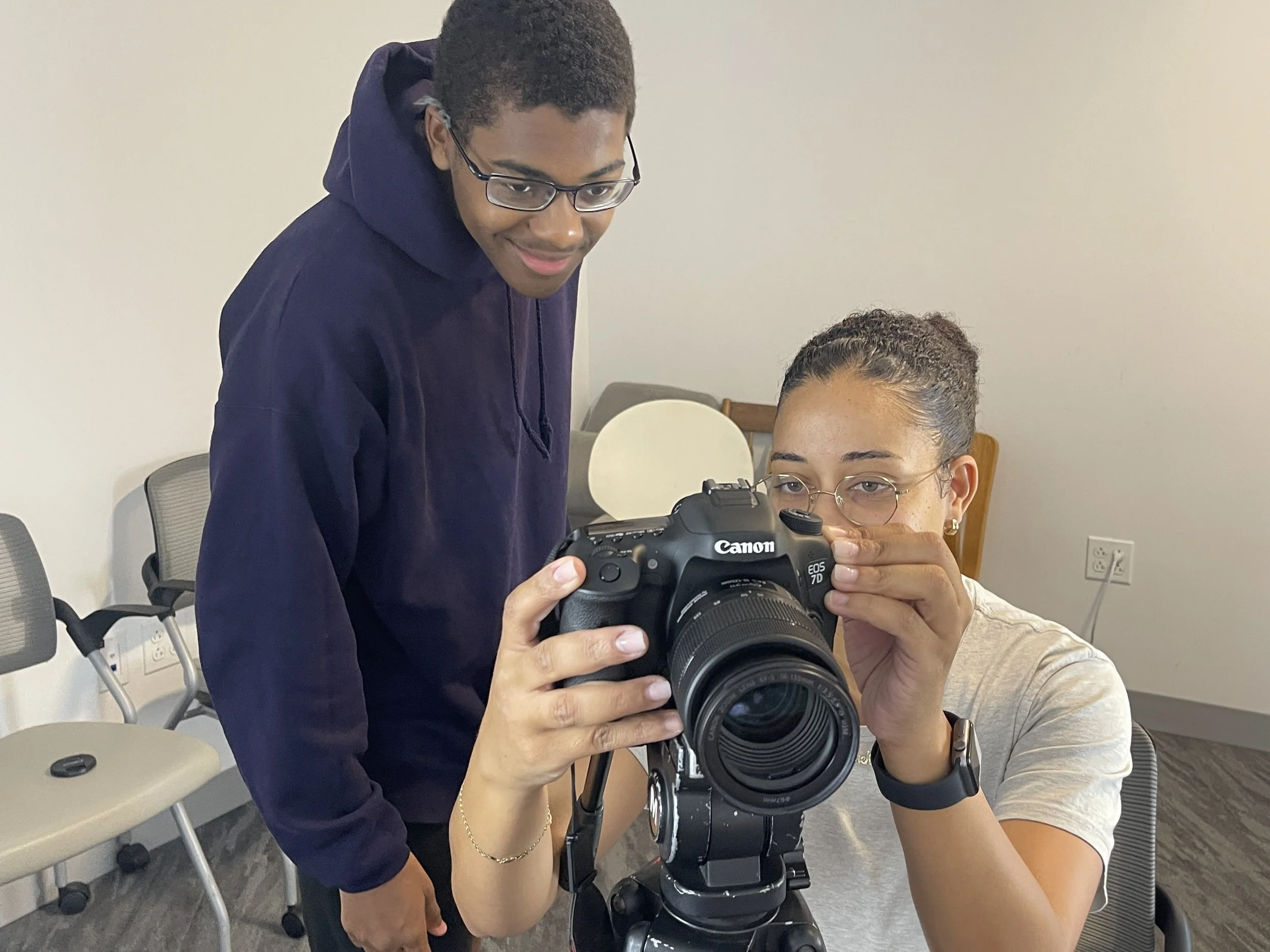 Two people. The person on the right is sitting down and operating a camera while the person on the left is stood and looking at the camera.