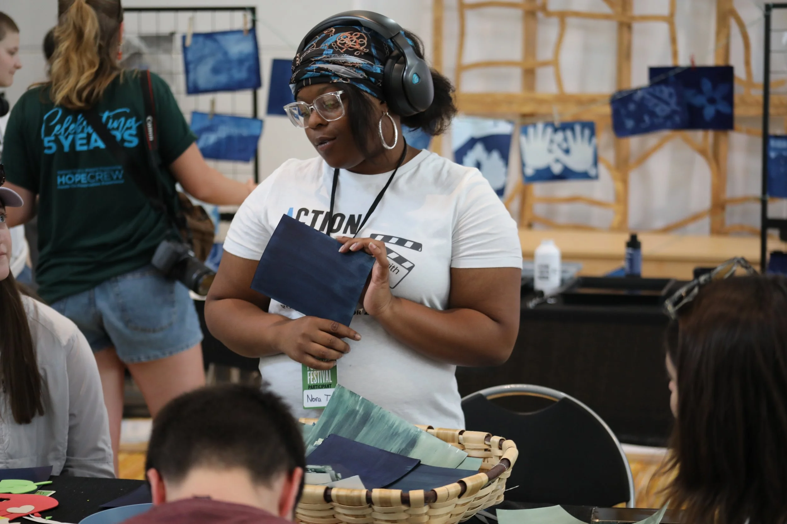 A person in an Action Youth Media shirt holding up a piece of painted paper used for cyanotype making. They appear to be explaining cyanotypes. Their name tag reads "Nora T."