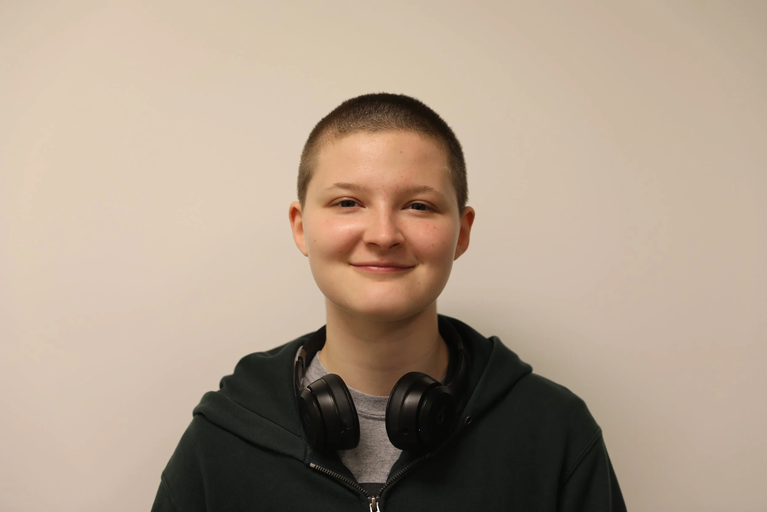 Light skinned person with a buzz cut, wearing a black zip-up hoodie, a gray tee shirt, and black headphones.