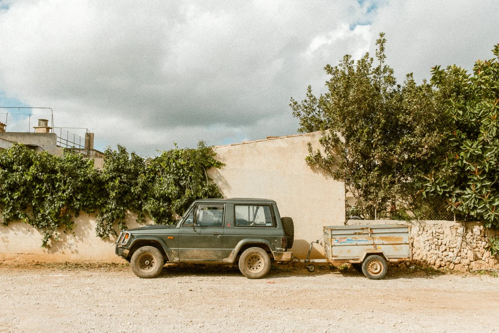 A black Jeep parked on a dirt street with a small trailer attached. The background features a beige wall, lush green bushes, and a cloudy sky.