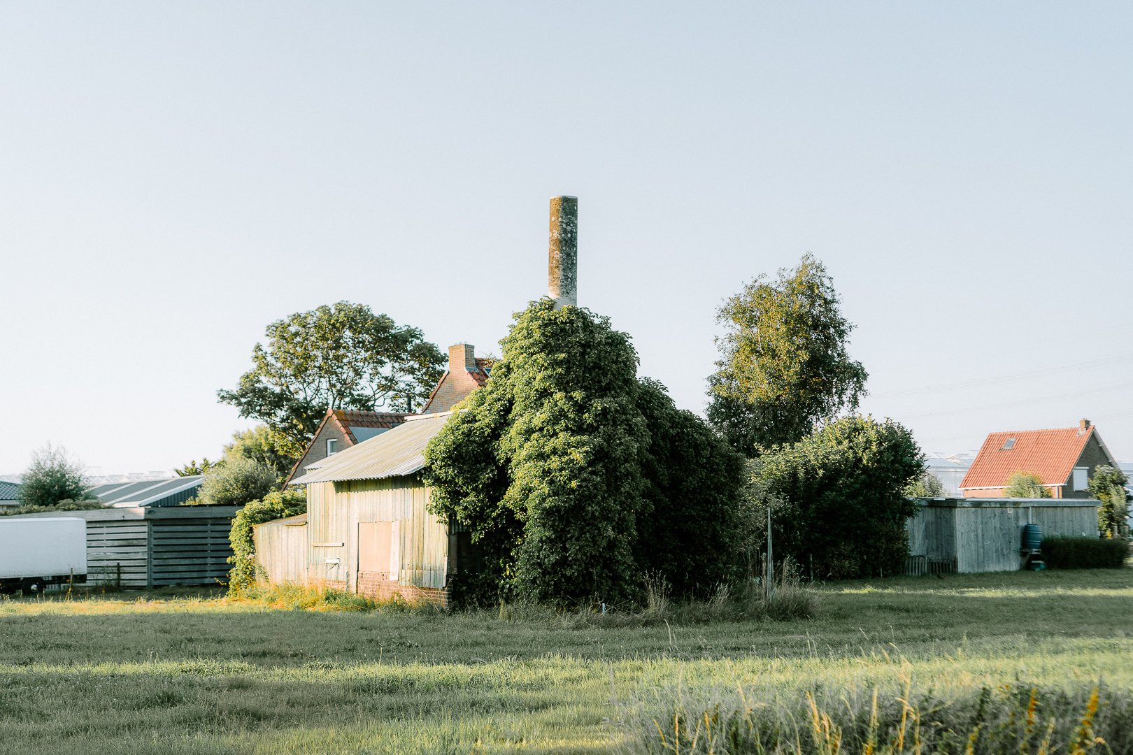 A rural scene with a weathered wooden barn surrounded by trees and grass, with houses and a tall smokestack in the background under a clear sky.