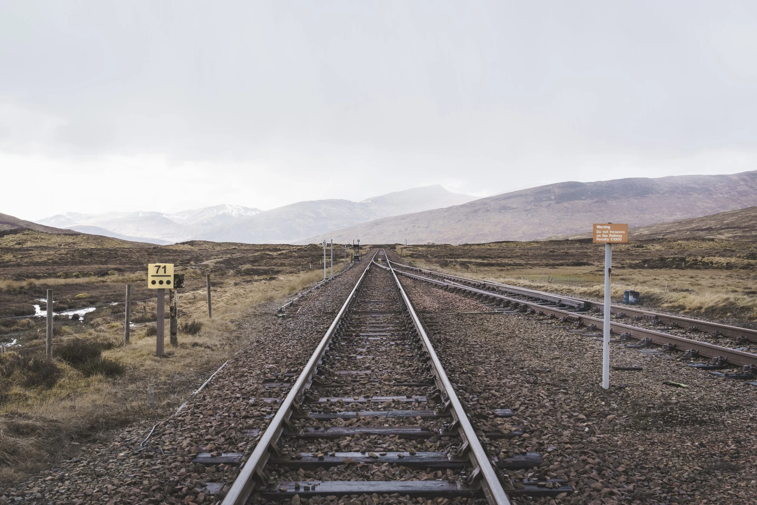 A railway track extends into the distance through a rural landscape with mountains in the background. There are warning signs near the tracks, one on the right and a yellow sign on the left. The sky is overcast.