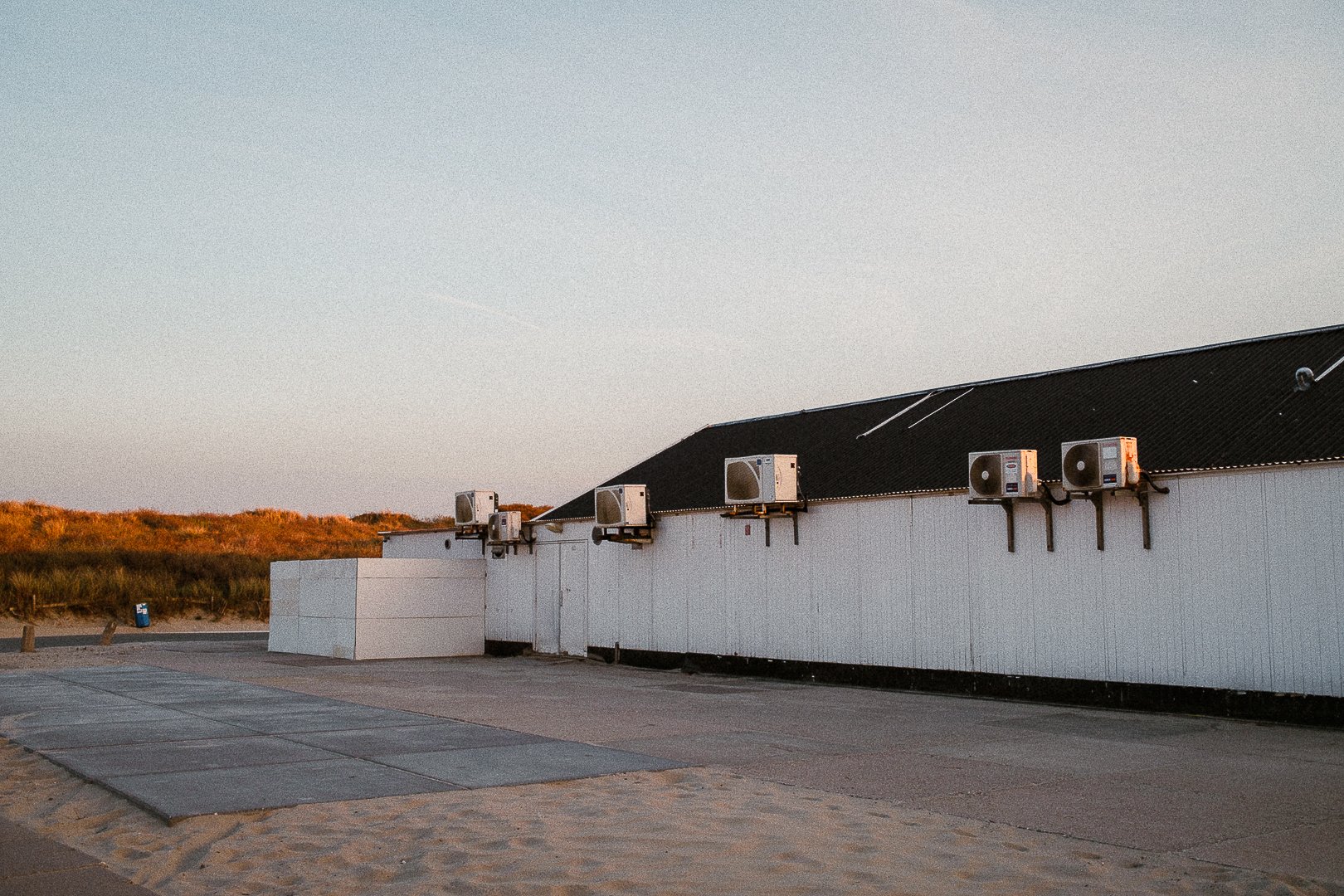 A building with a dark sloped roof and several outdoor air conditioning units mounted on the white wall, with a sandy area and grassy dunes in the background during sunset.
