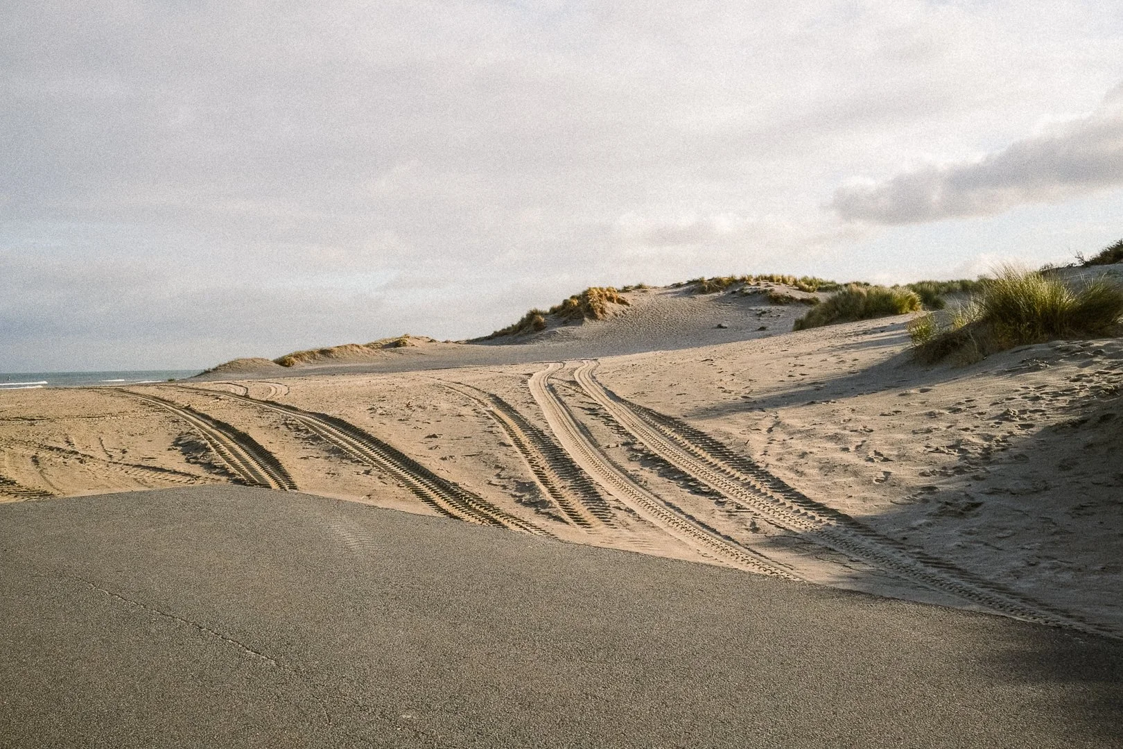 A sandy beach with tire tracks and patches of grass, with dunes and cloudy sky in the background.