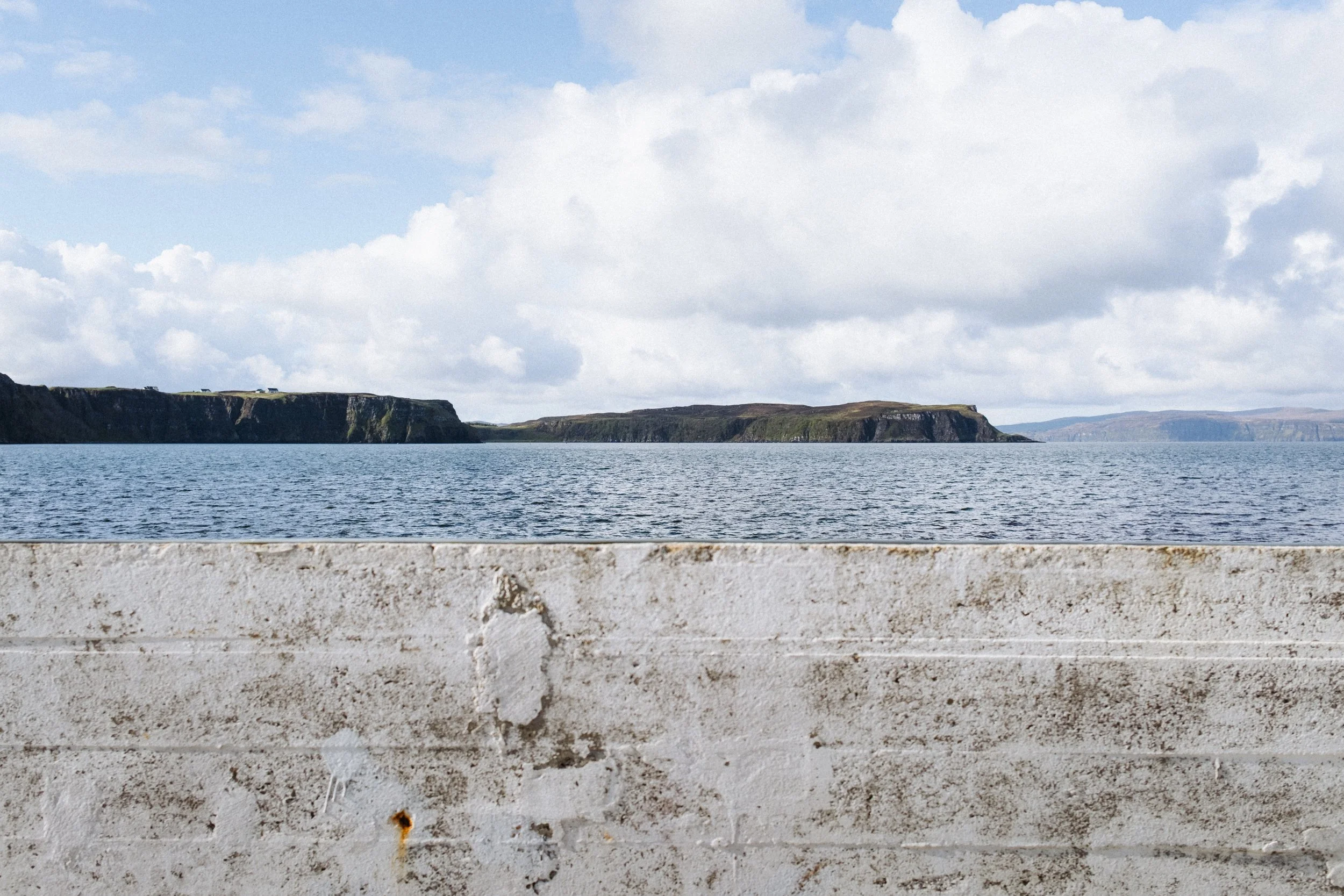View of a body of water with cliffs and a partly cloudy sky in the background, and a textured stone or concrete surface in the foreground.