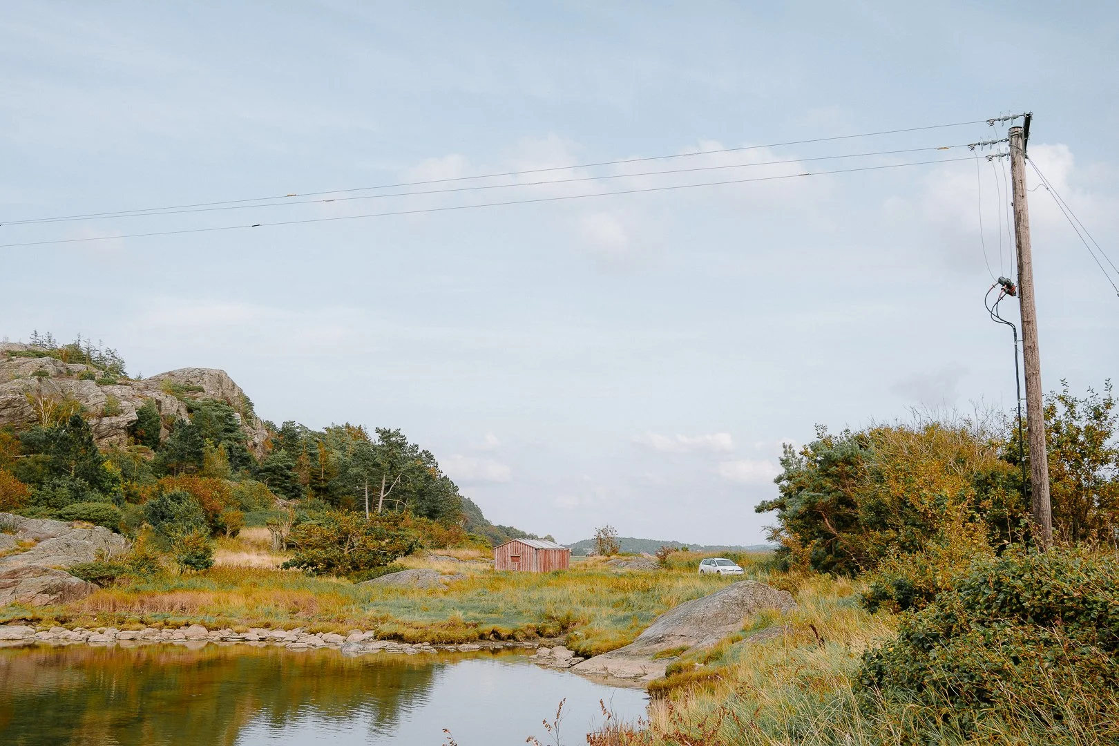 A rural landscape with a creek in the foreground, a grassy field, a small wooden building, trees, a utility pole, and a car in the distance under a partly cloudy sky.