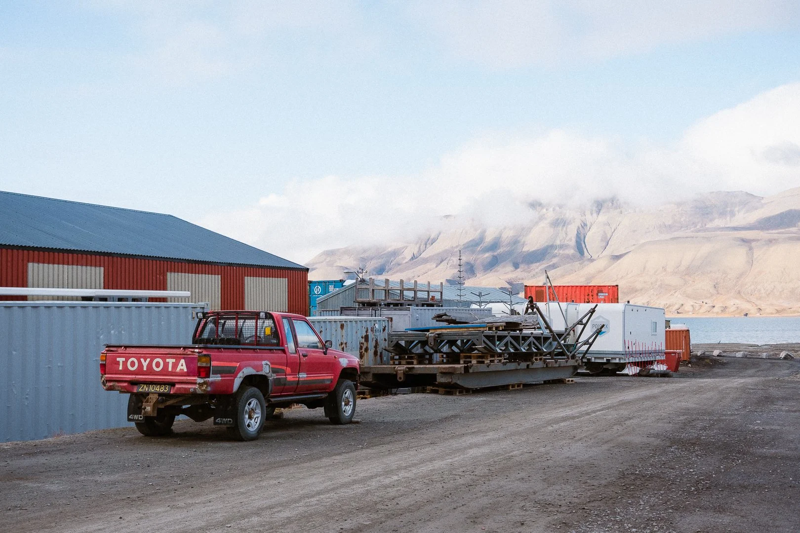 A red Toyota pickup truck parked on a dirt surface near cargo containers, with a large red building, mountains, and water in the background.