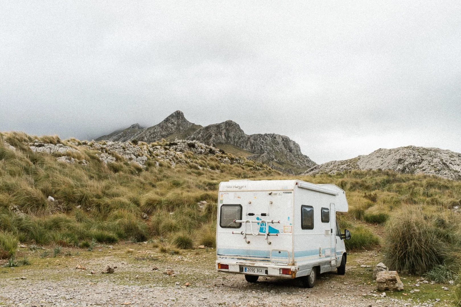 A white camper van parked on a dirt clearing surrounded by grassy hills and rocky mountains under an overcast sky.