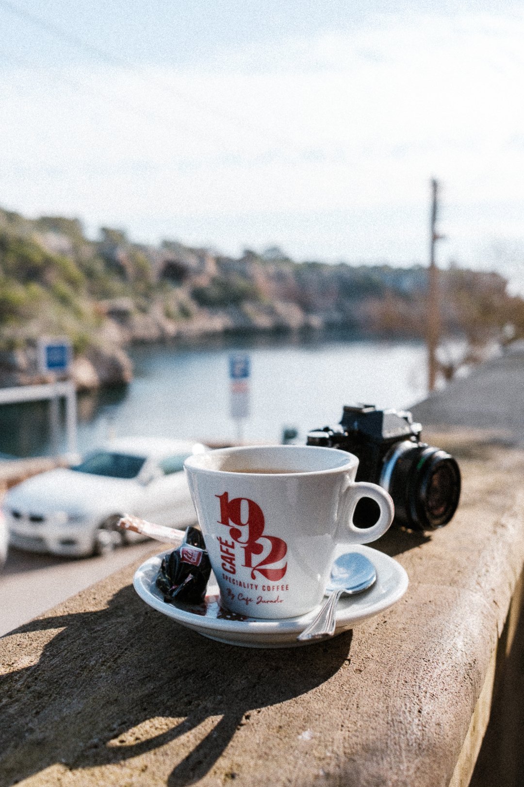 A cup of coffee and an analog camera standing in the sunny spot with a bay in the background