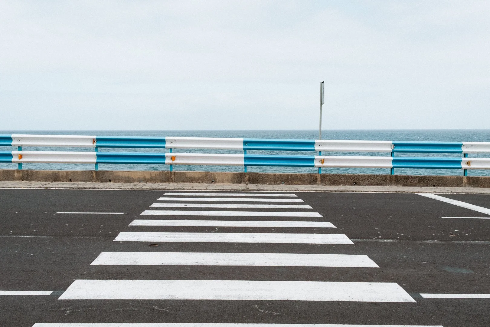 A crosswalk on a road next to a blue and white guardrail, with the ocean visible in the background under an overcast sky.