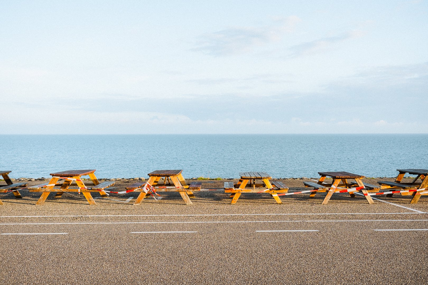 Empty wooden picnic tables along a waterfront with a body of water and cloudy sky in the background.