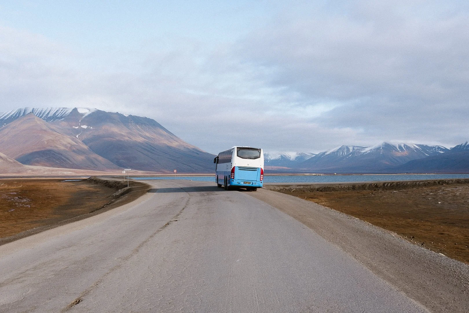 A small blue and white bus driving on a unpaved road in a scenic landscape with mountains and a body of water in the background.
