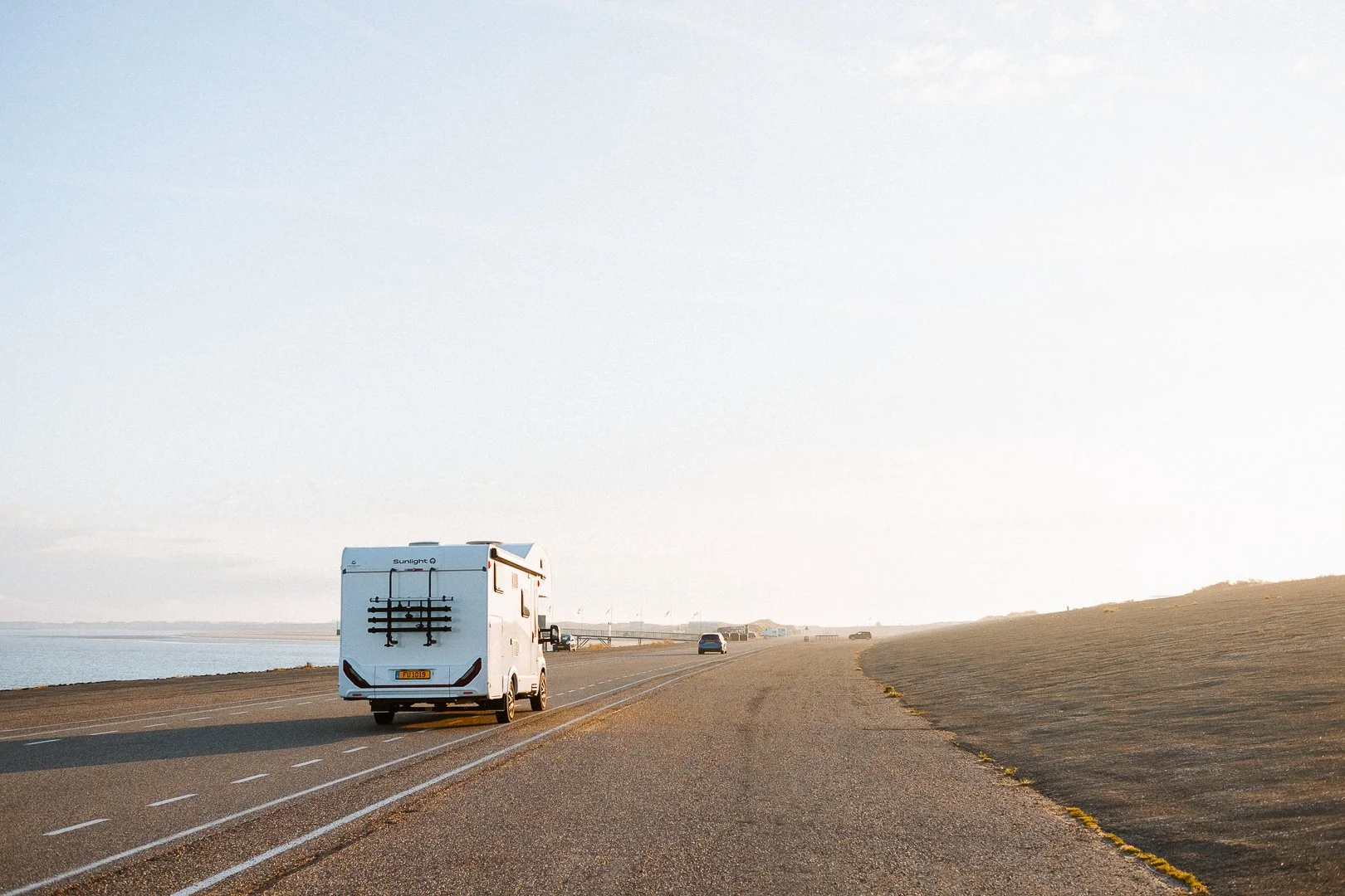 A recreational vehicle travels along a coastal road during late afternoon or early evening, with the ocean on the left and a sloped hill on the right, under a clear sky.