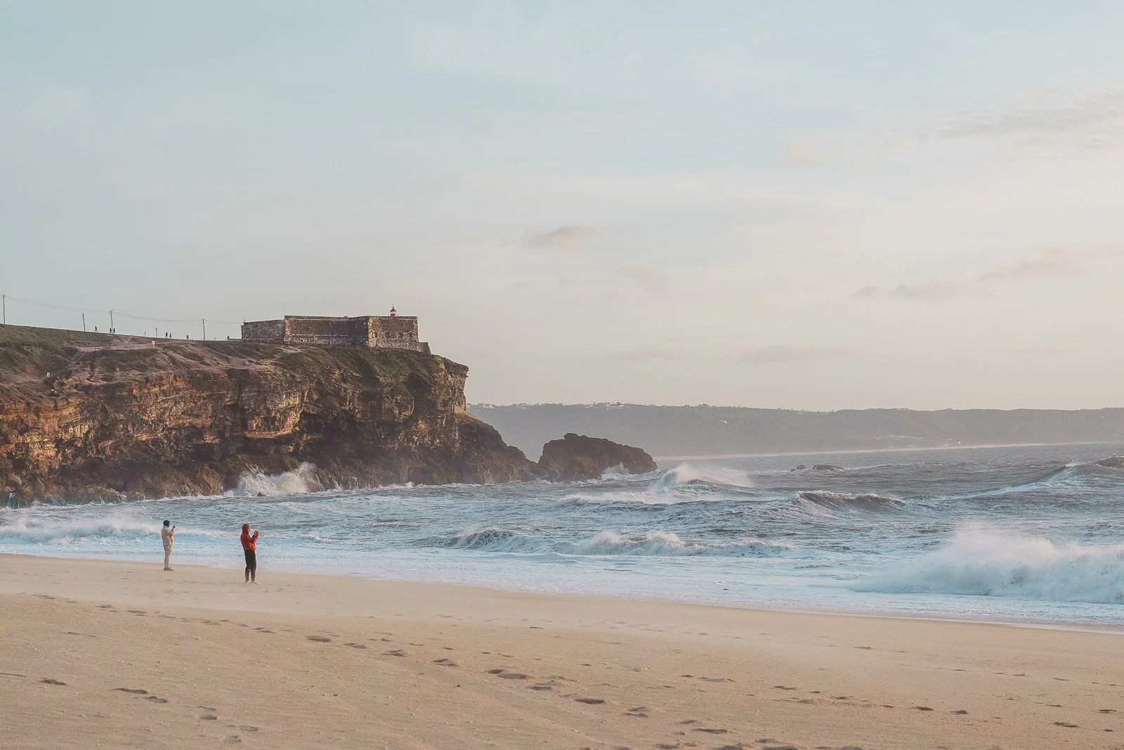 A beach scene with two people taking photos and footprints in the sand. There are waves crashing on the shore, a rocky cliff with a historic building on top, and a cloudy sky.