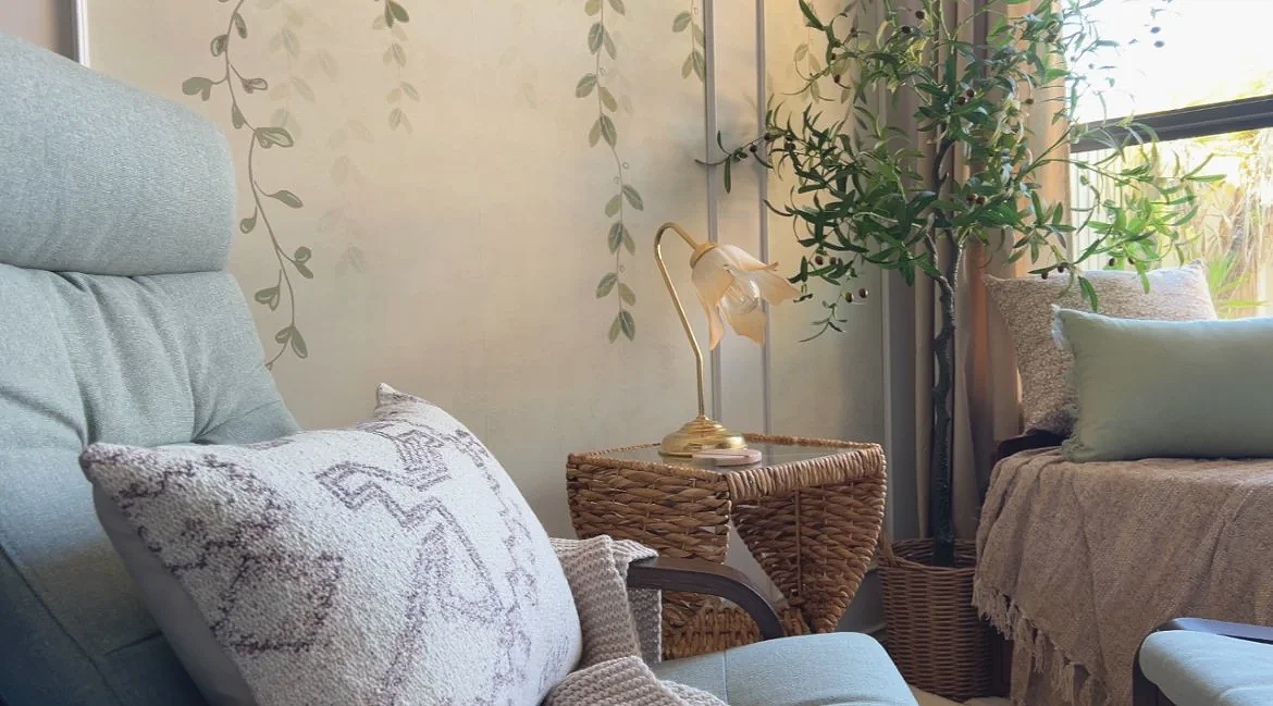 Cozy living room corner with a light blue armchair, patterned pillows, a wicker side table with a gold desk lamp, a large potted plant, and window with natural light.