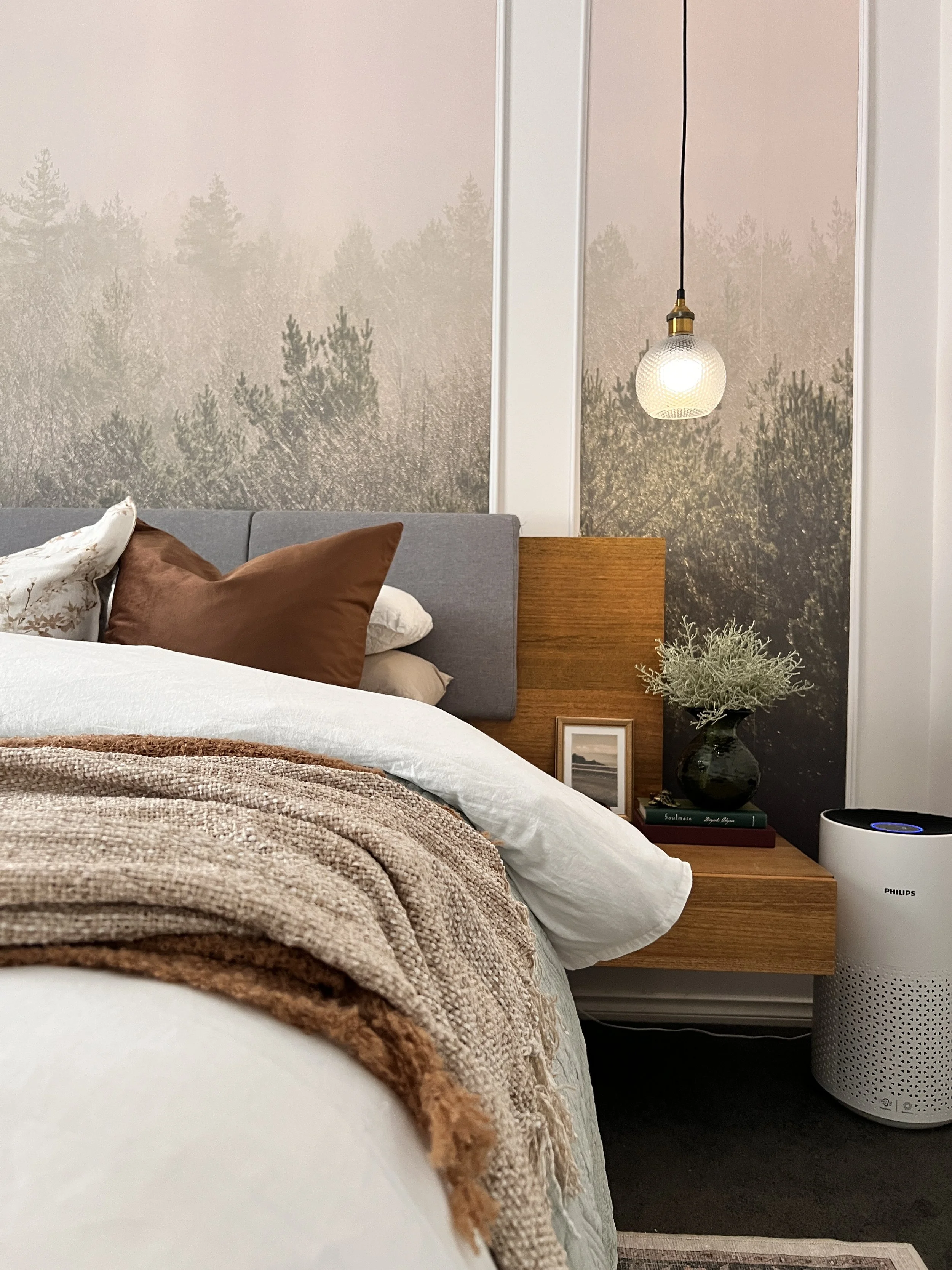 Bedroom with a bed featuring white, brown, and patterned pillows, a textured beige blanket, a wooden nightstand with books, a plant in a black vase, a framed picture, and a hanging pendant light, with a forest wallpaper and an air purifier.