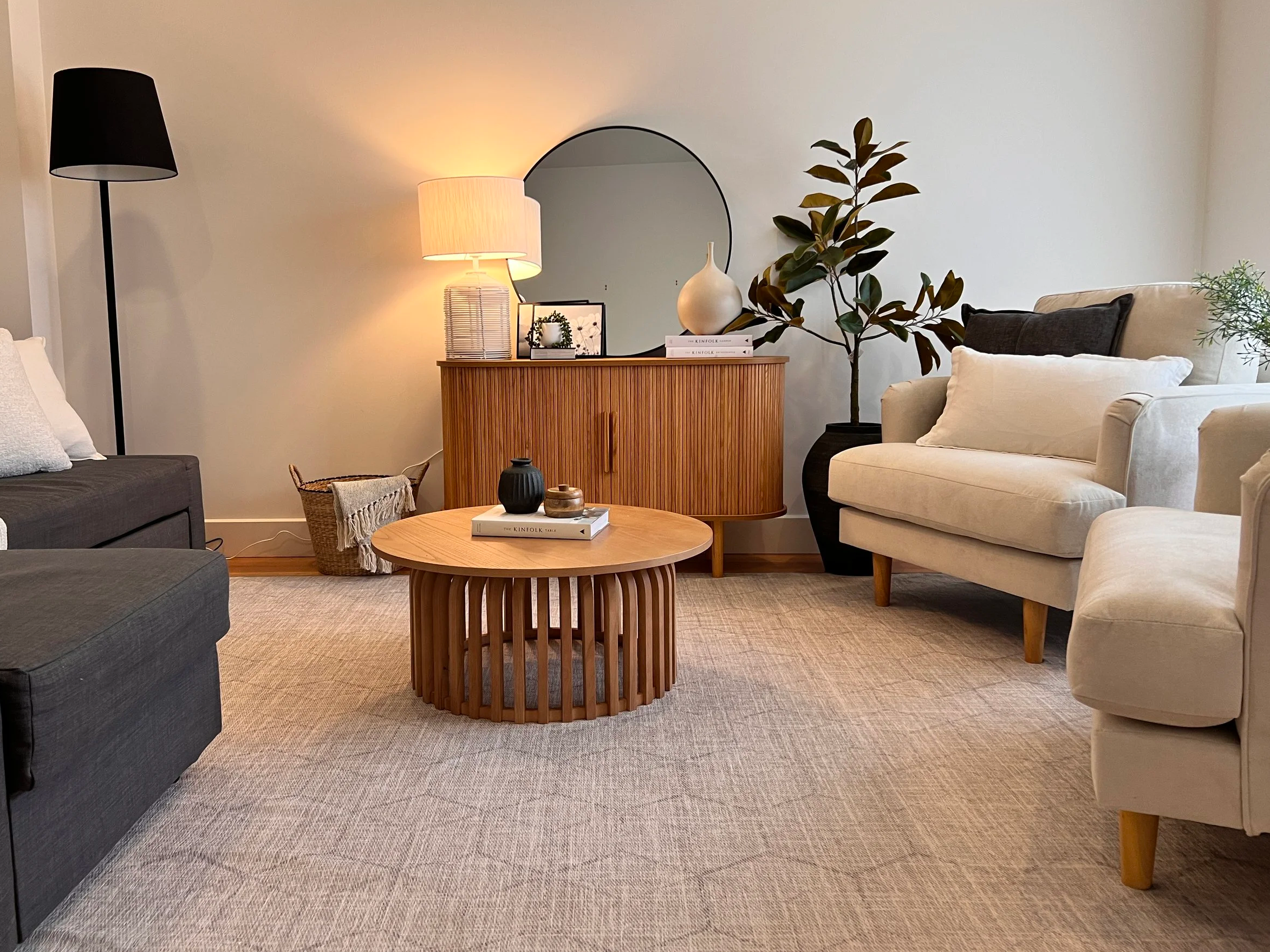 Living room with beige carpet, two white armchairs, a dark gray sofa, a wooden round coffee table with decorative objects, a wooden sideboard with a mirror, lamp, and plants, and various decorative items and framed pictures.