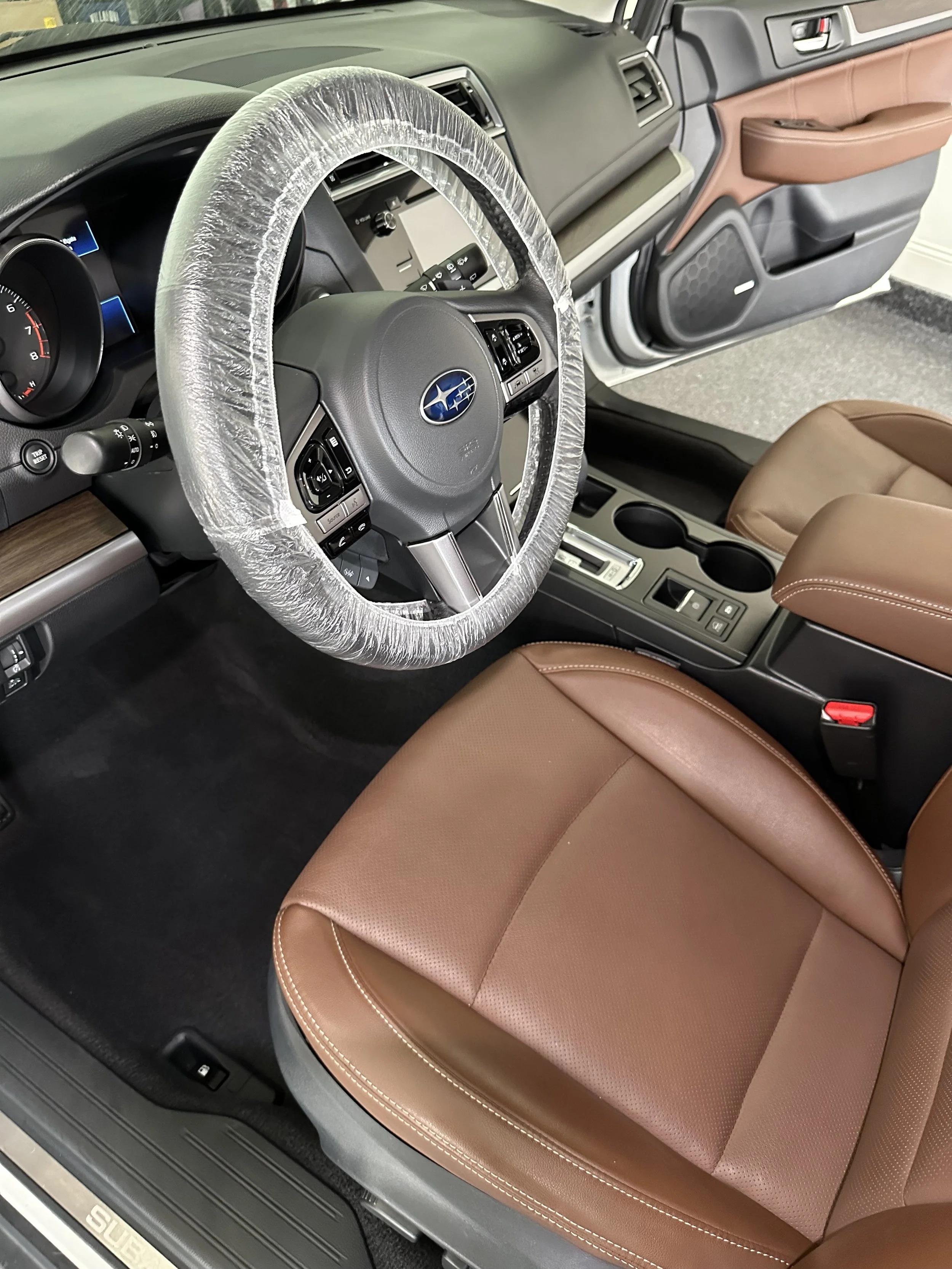Interior of a vehicle showing the steering wheel with a protective plastic cover, brown leather seats, and part of the dashboard with control buttons and cup holders.