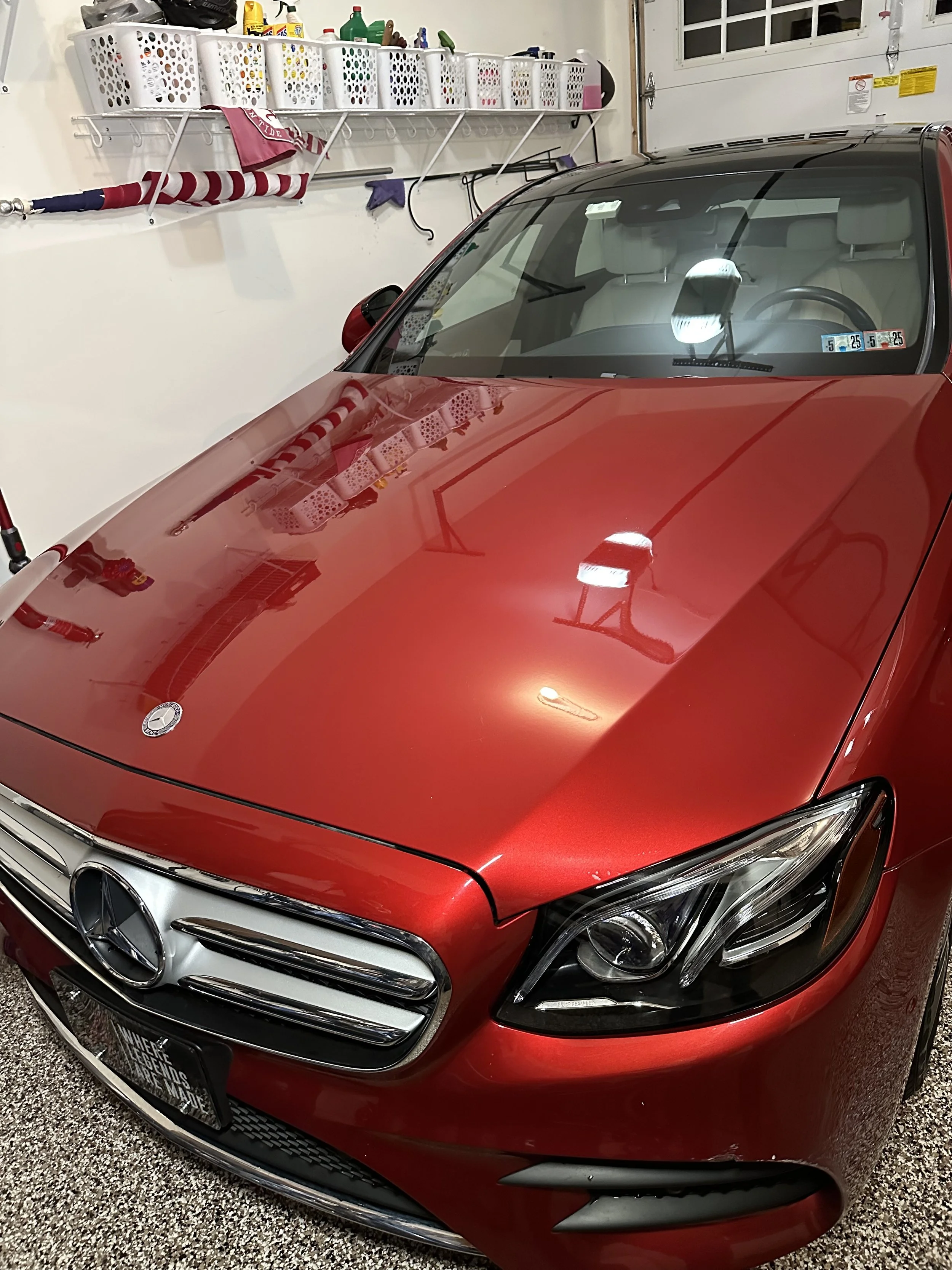A red Mercedes-Benz car parked in a garage with a white wall. The garage has shelves with various cleaning supplies and laundry items. The car's hood and front are visible, with reflections of the garage lights.