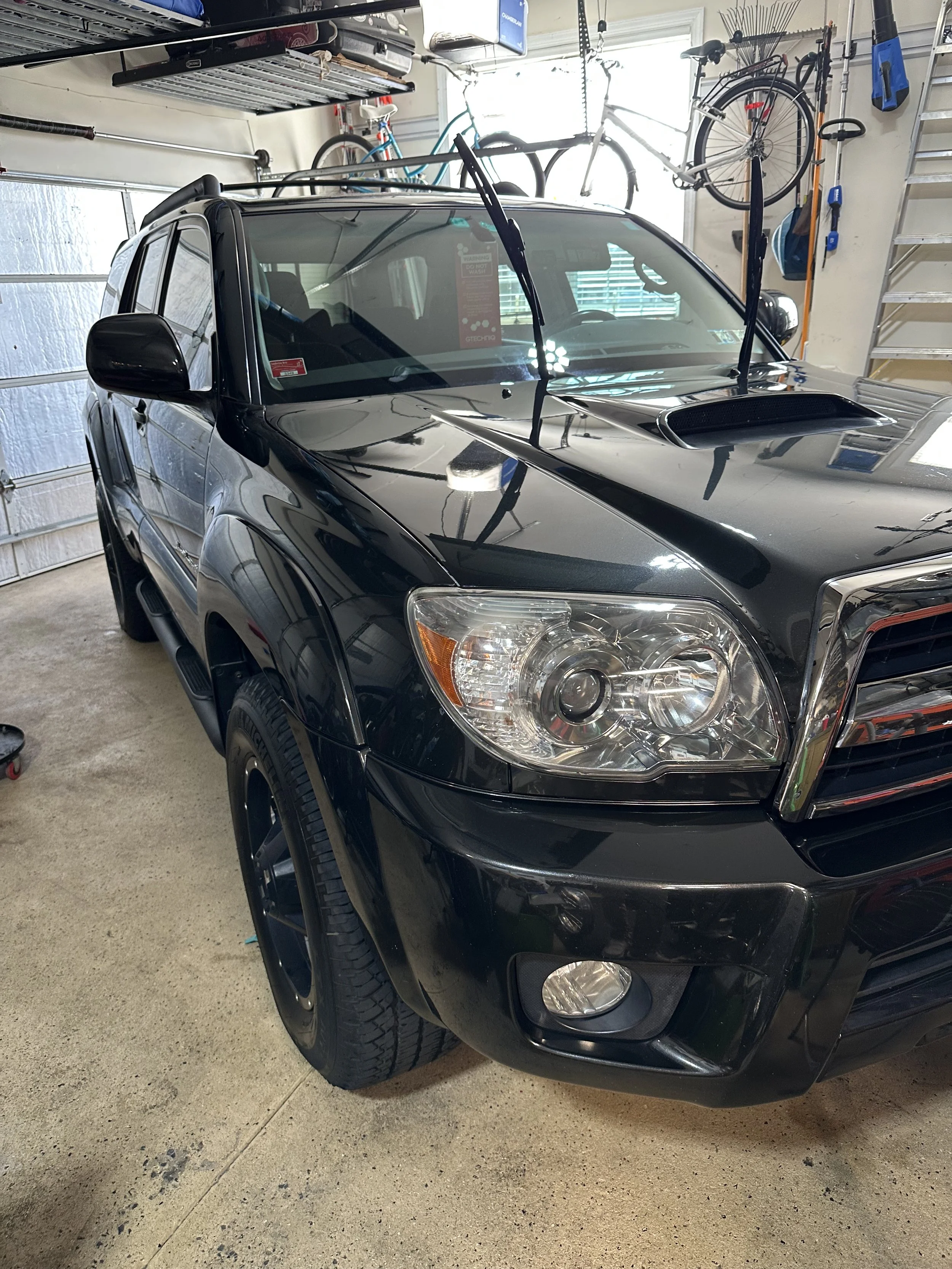 Black SUV parked in a garage with bicycles hanging on the wall above. Garage wall and tools visible in the background.