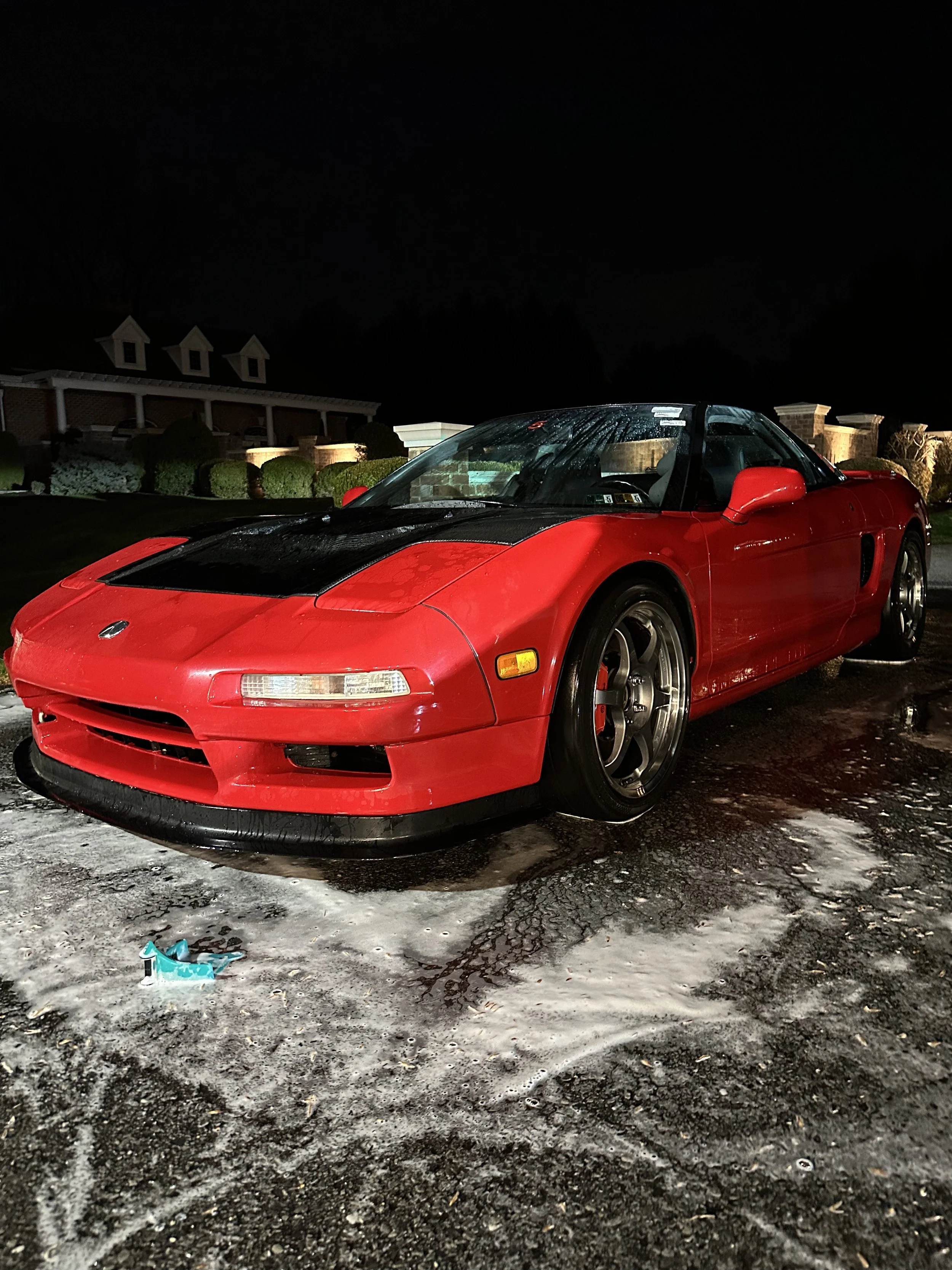 Red sports car parked outdoors at night, with soap and water on the ground indicating it was recently washed.