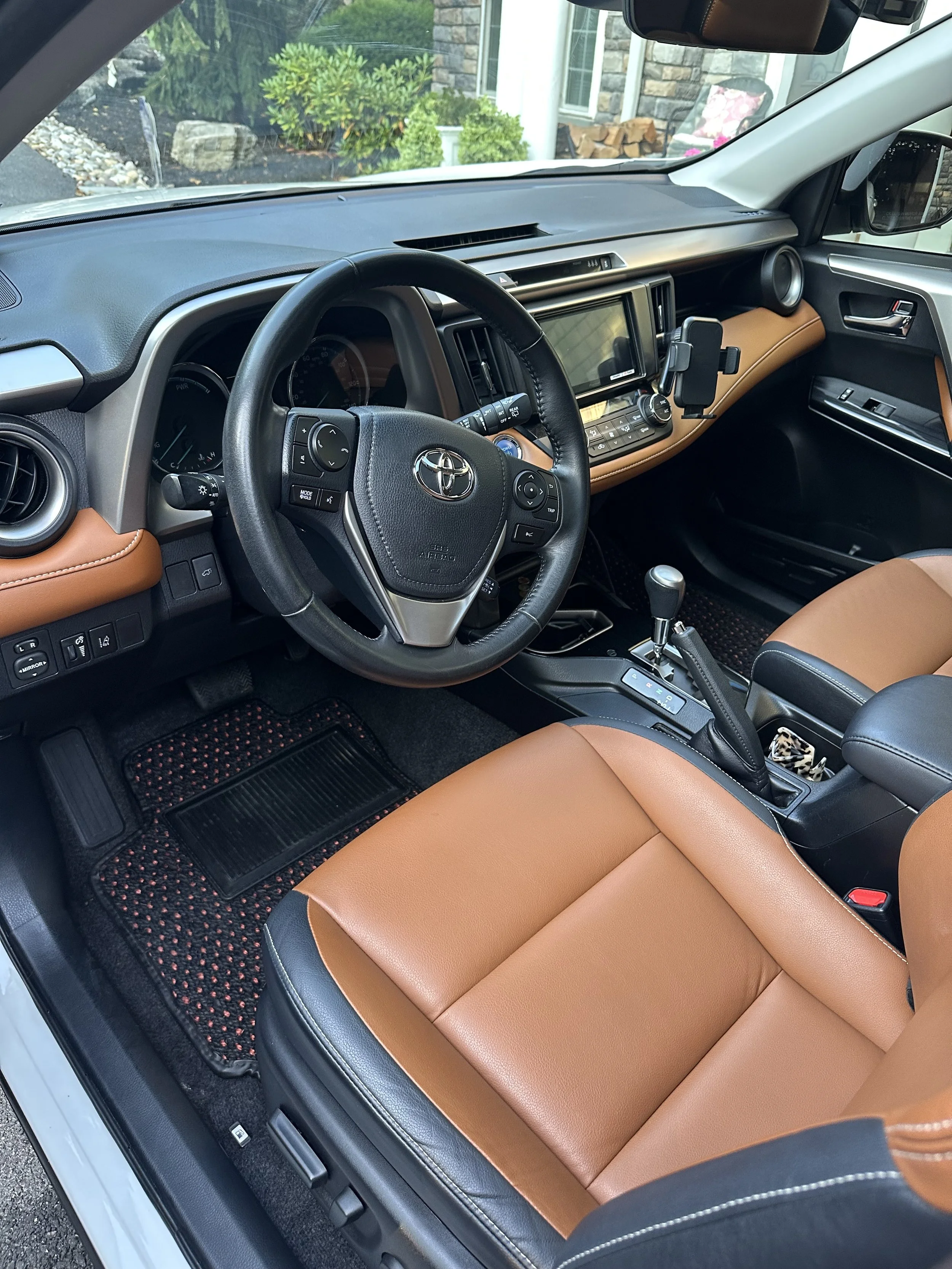 Interior view of a Toyota vehicle showing a black and tan dashboard, steering wheel, gear shift, and tan leather seats. There is an Android phone mounted on the dashboard.