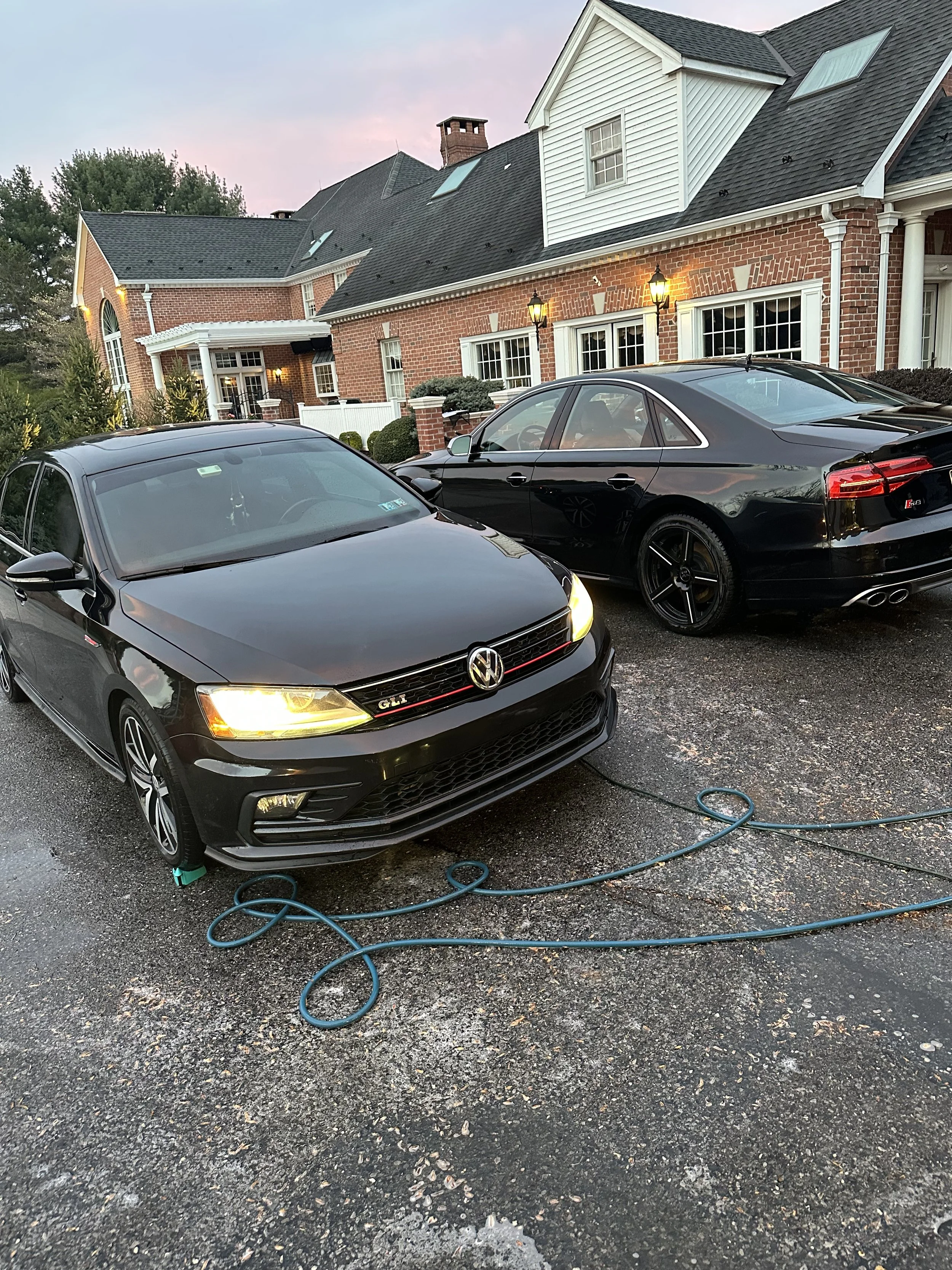 Two black cars, a Volkswagen and an Audi, parked in front of a large brick house. The Volkswagen's headlights are on, and a garden hose is lying on the wet asphalt driveway.