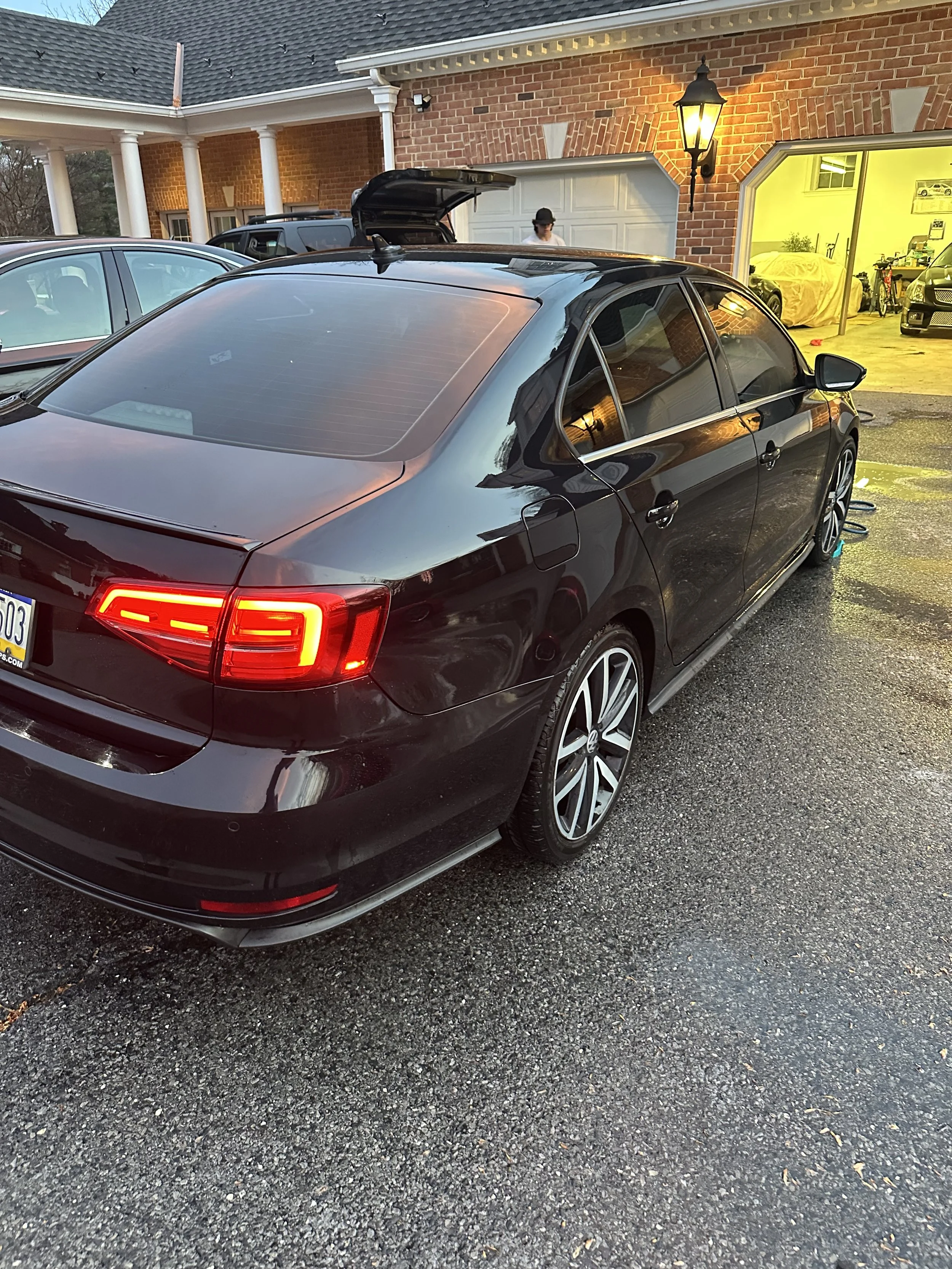 Black hatchback car parked in driveway near house with brick wall, garage, and illuminated lamp post, with a person and other cars nearby, and a brightly lit garage interior.