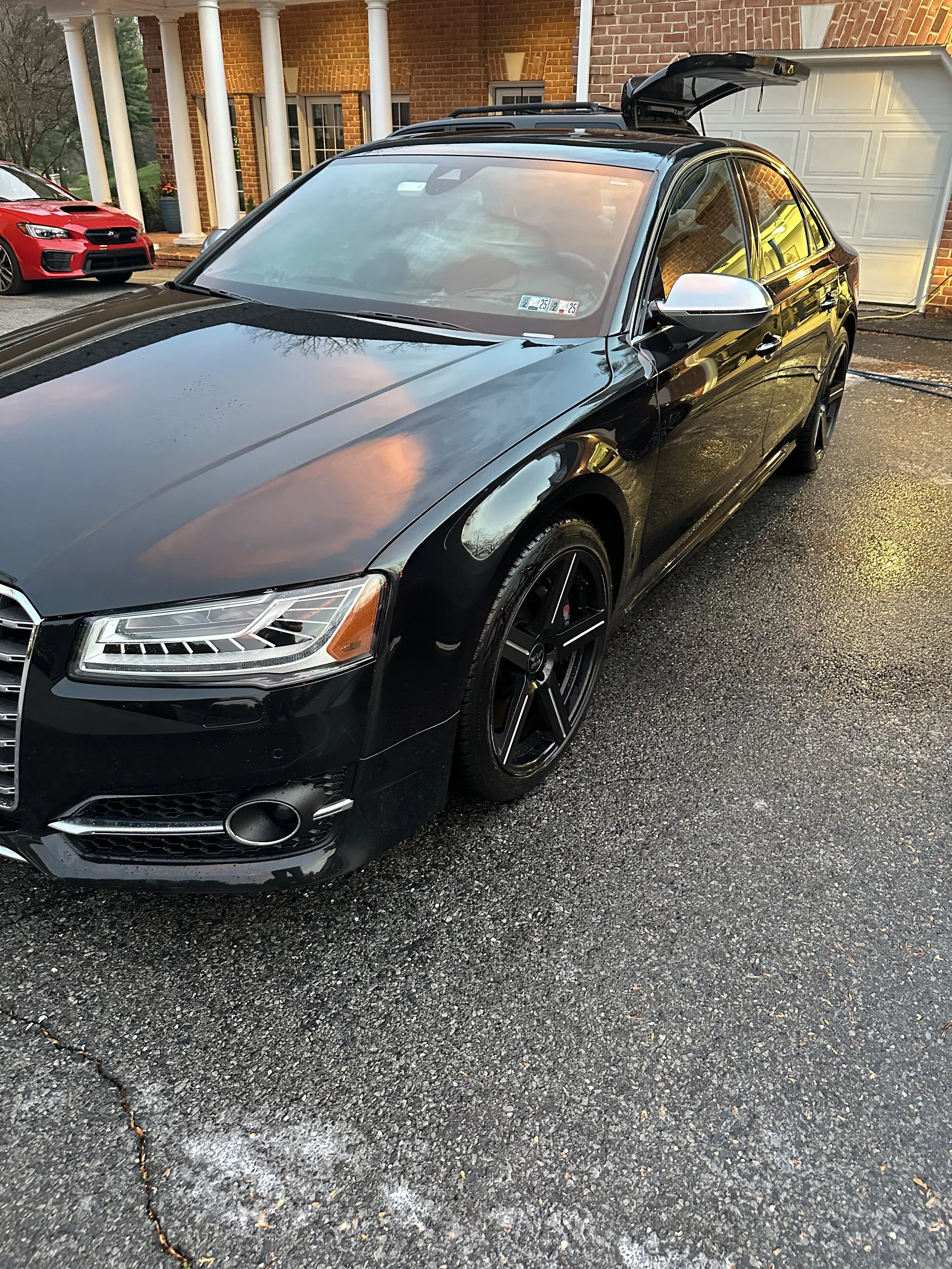 A black Mercedes-Benz sedan with tinted windows and silver side mirrors parked in a driveway. The car has black rims and is wet, reflecting the sky and surroundings.