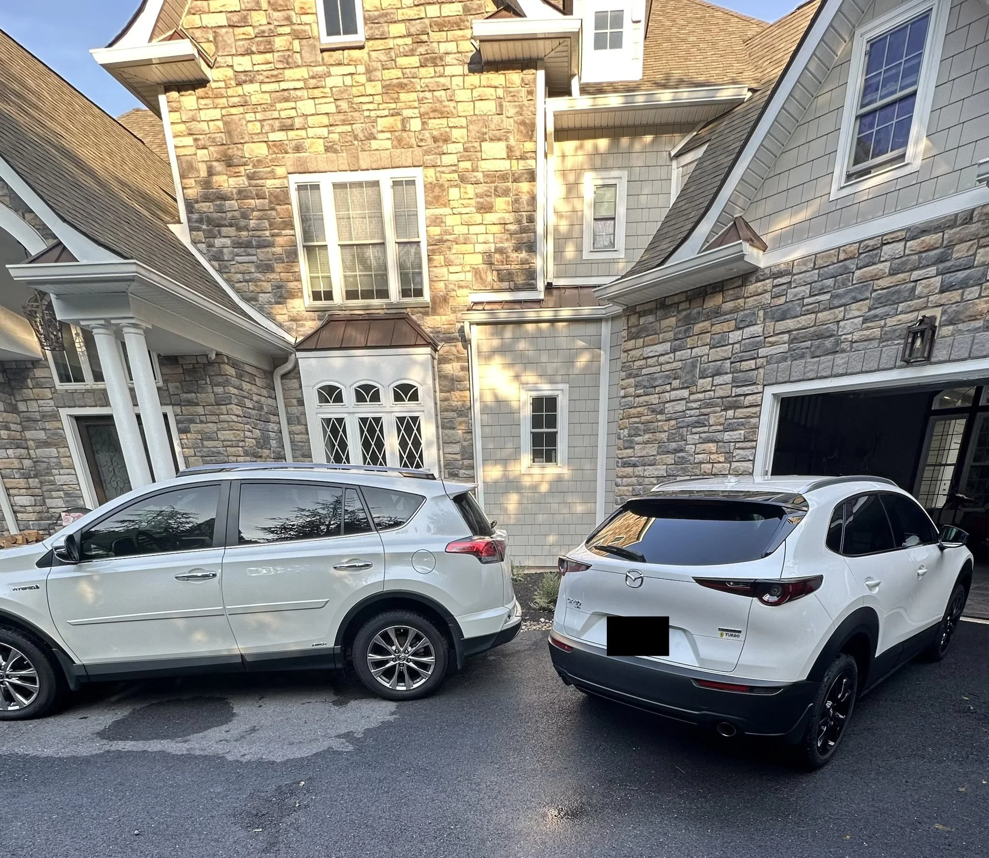 Two white SUVs parked in front of a large, multi-story house with stone and siding exterior. The house has multiple windows, some with decorative glass, and features a walkway with columns near the entrance.