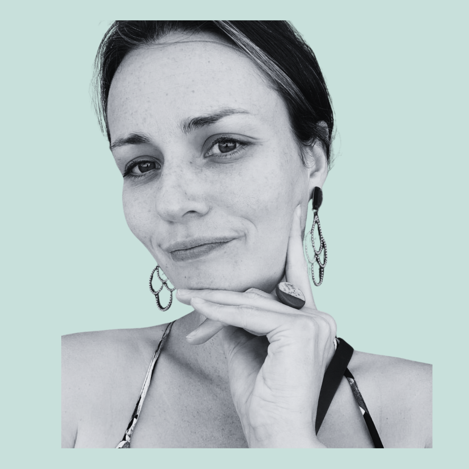 Close-up of a woman with short dark hair, wearing earrings, a ring, and a tank top, posing with her hand on her chin against a plain background.