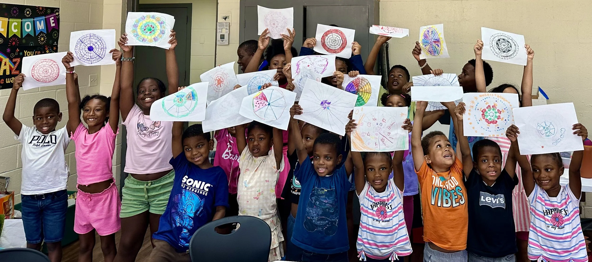 A large group of smiling children holding their hand-drawn colorful mandala artwork over their heads at a Sumac Arts community workshop for kids