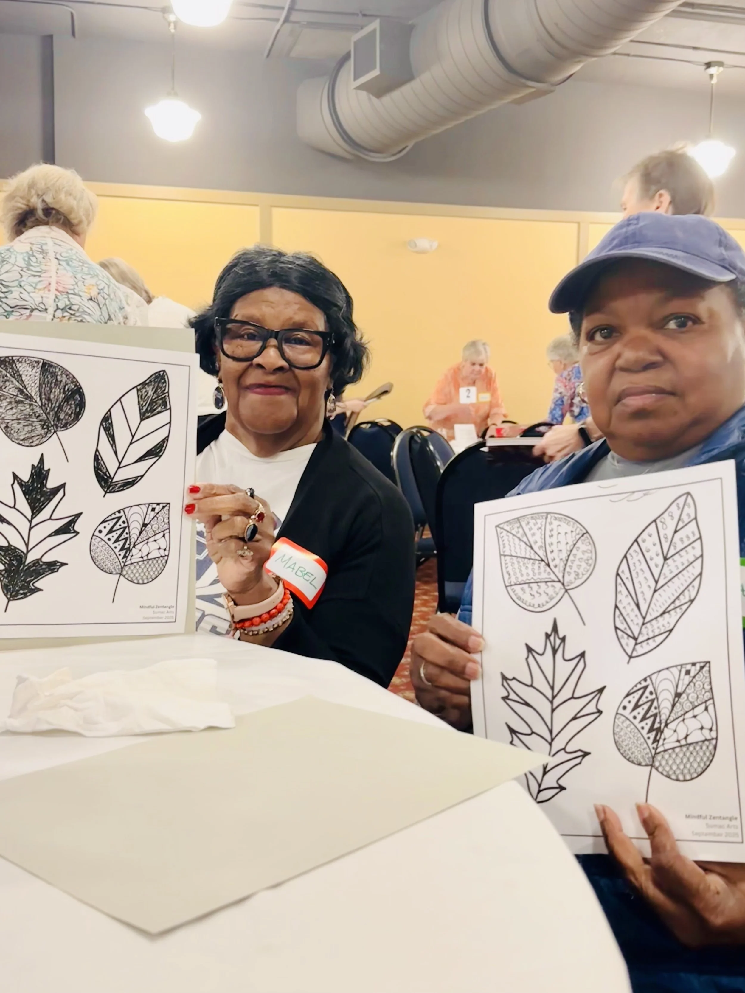 Two senior women smiling and showcasing their mindful hand-drawn leaf artwork at a Sumac Arts presentation in Newberry, South Carolina